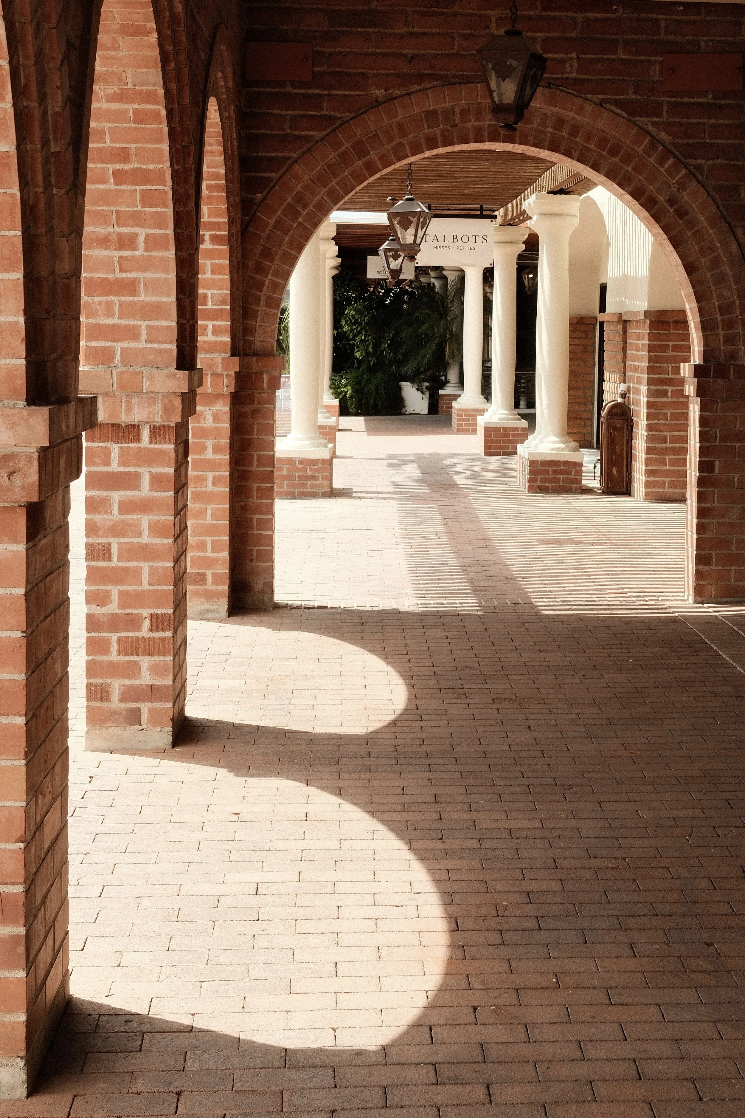 A brick walkway under archways with hanging lanterns and white columns, leading to a sign that reads 'Balbots Misses Petites' in the background, with greenery and sunlight casting shadows.
