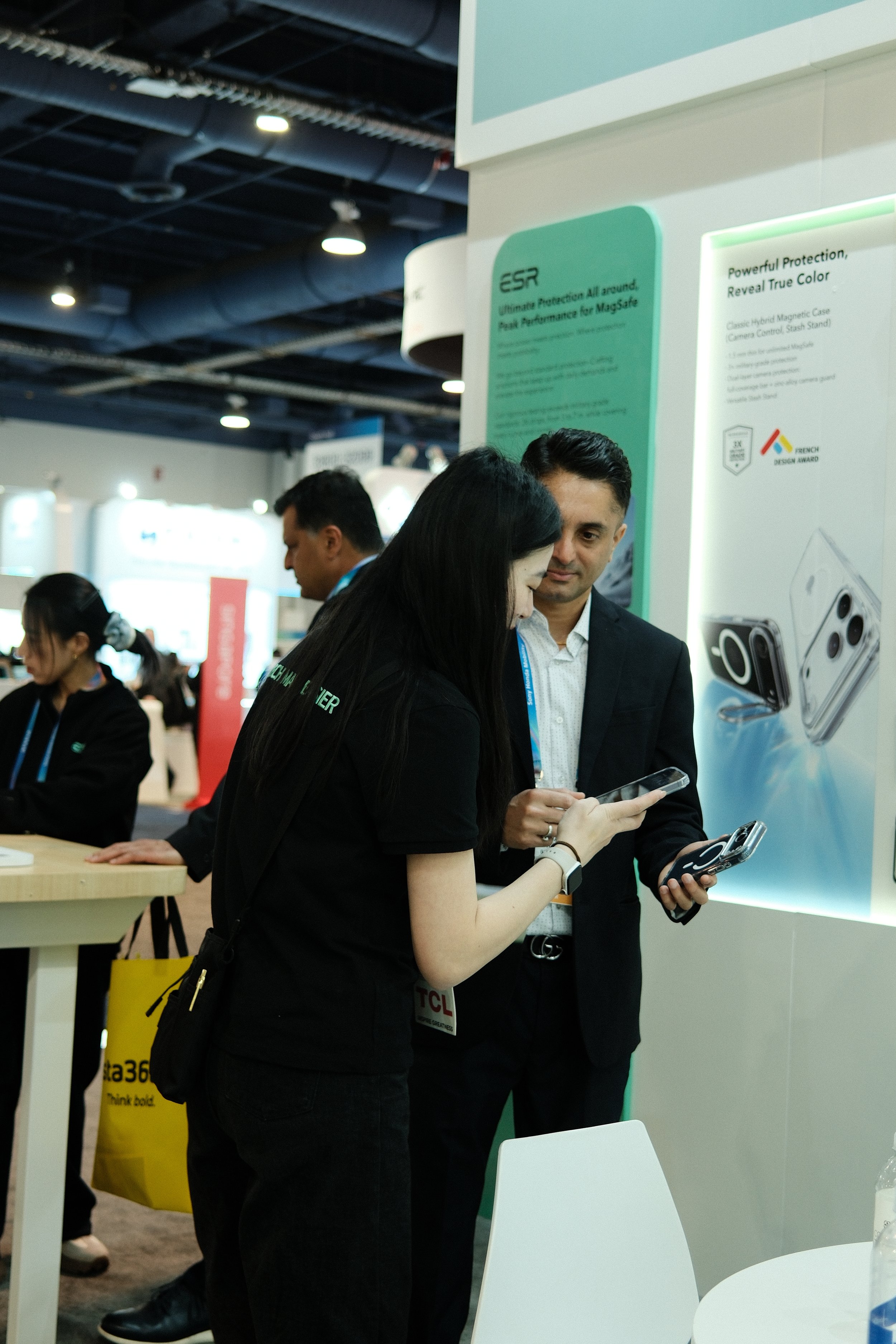 Two people, a woman and a man, standing together at a trade show booth, looking at their smartphones. The booth has a display with images of smartphones and text about protection cases.