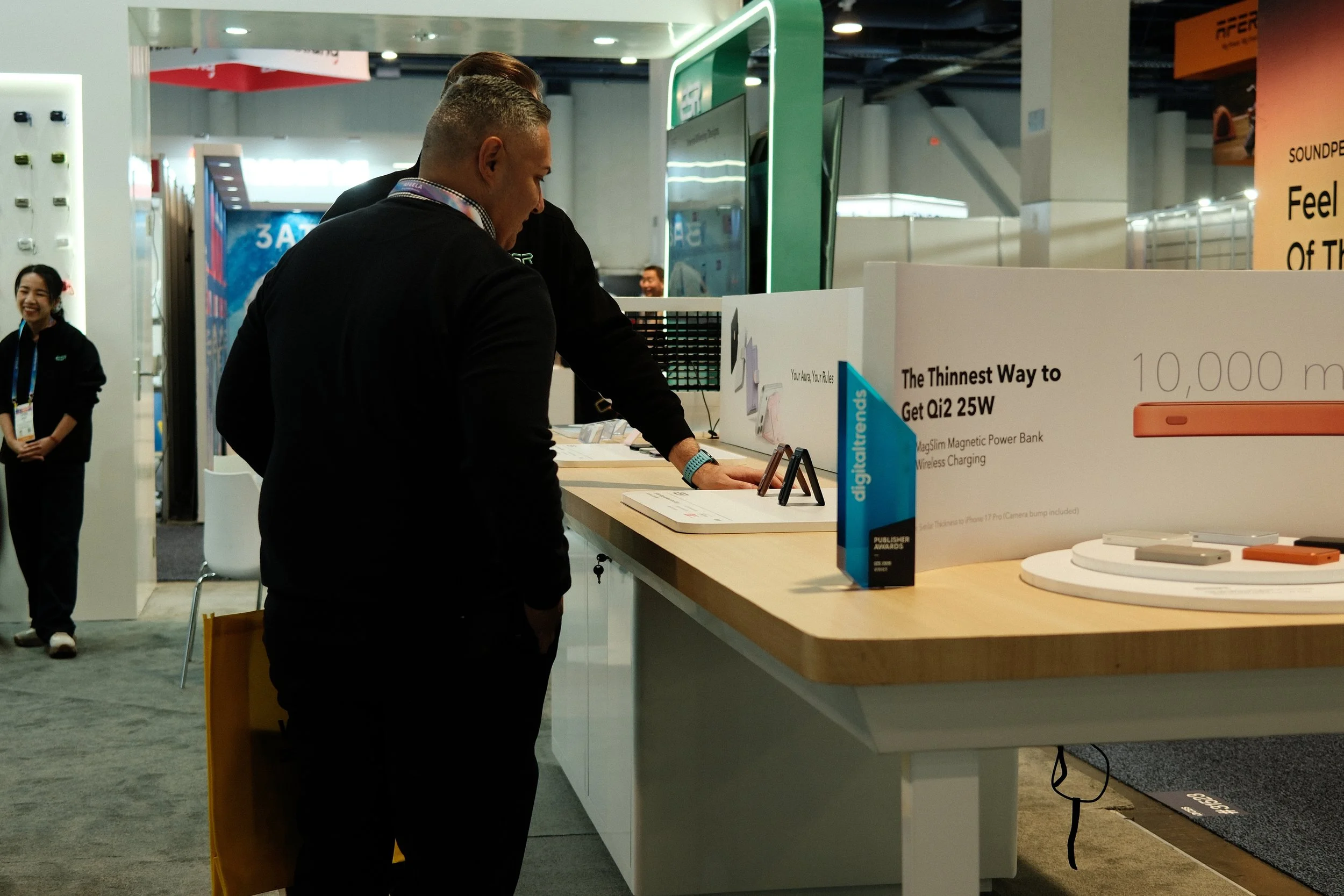 A man and a woman are at a technology booth showcasing electronic gadgets, with the man leaning over the counter to look at items and the woman smiling in the background.