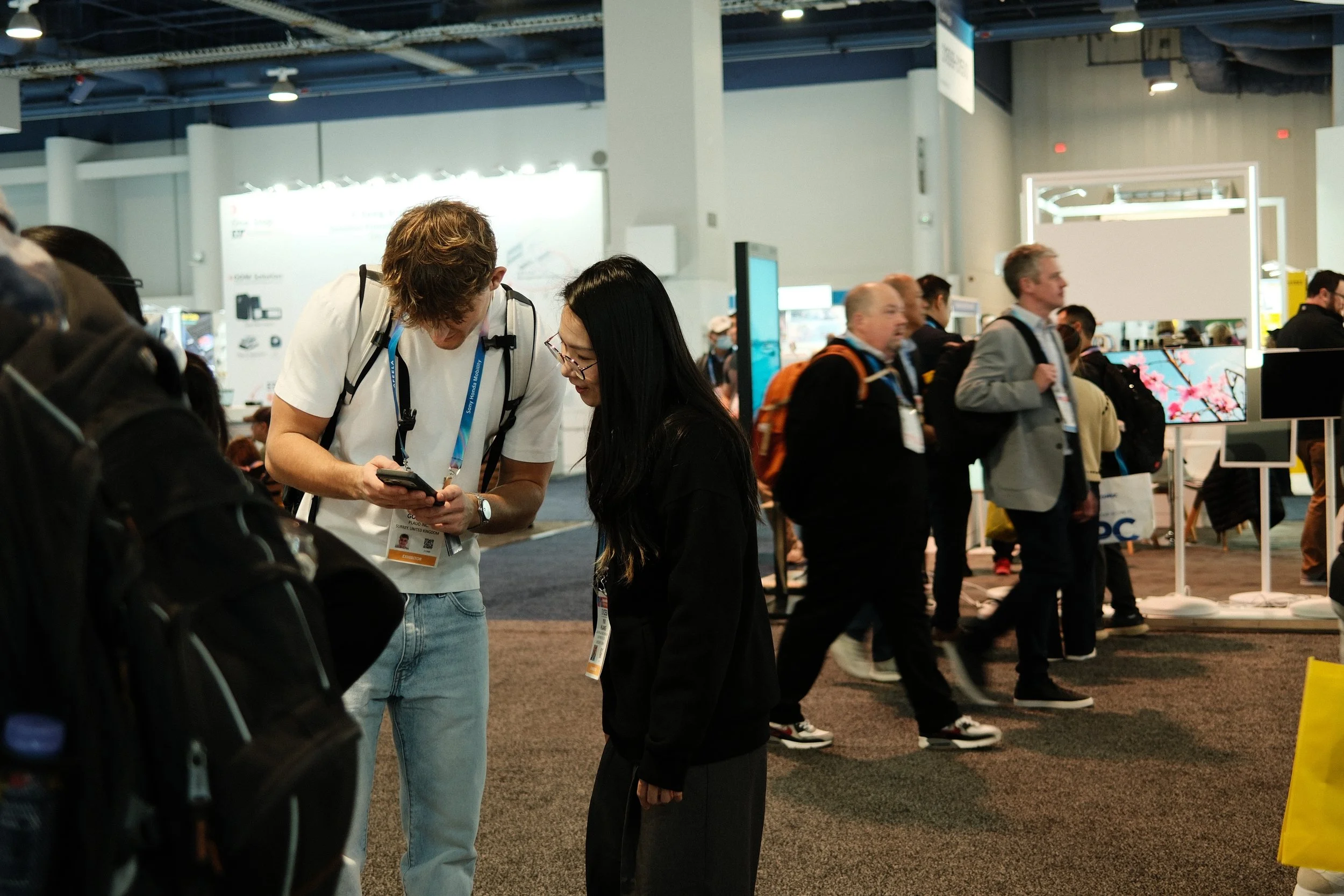 Two people are looking at a smartphone at a conference or trade show, with several other attendees walking by and displays visible in the background.
