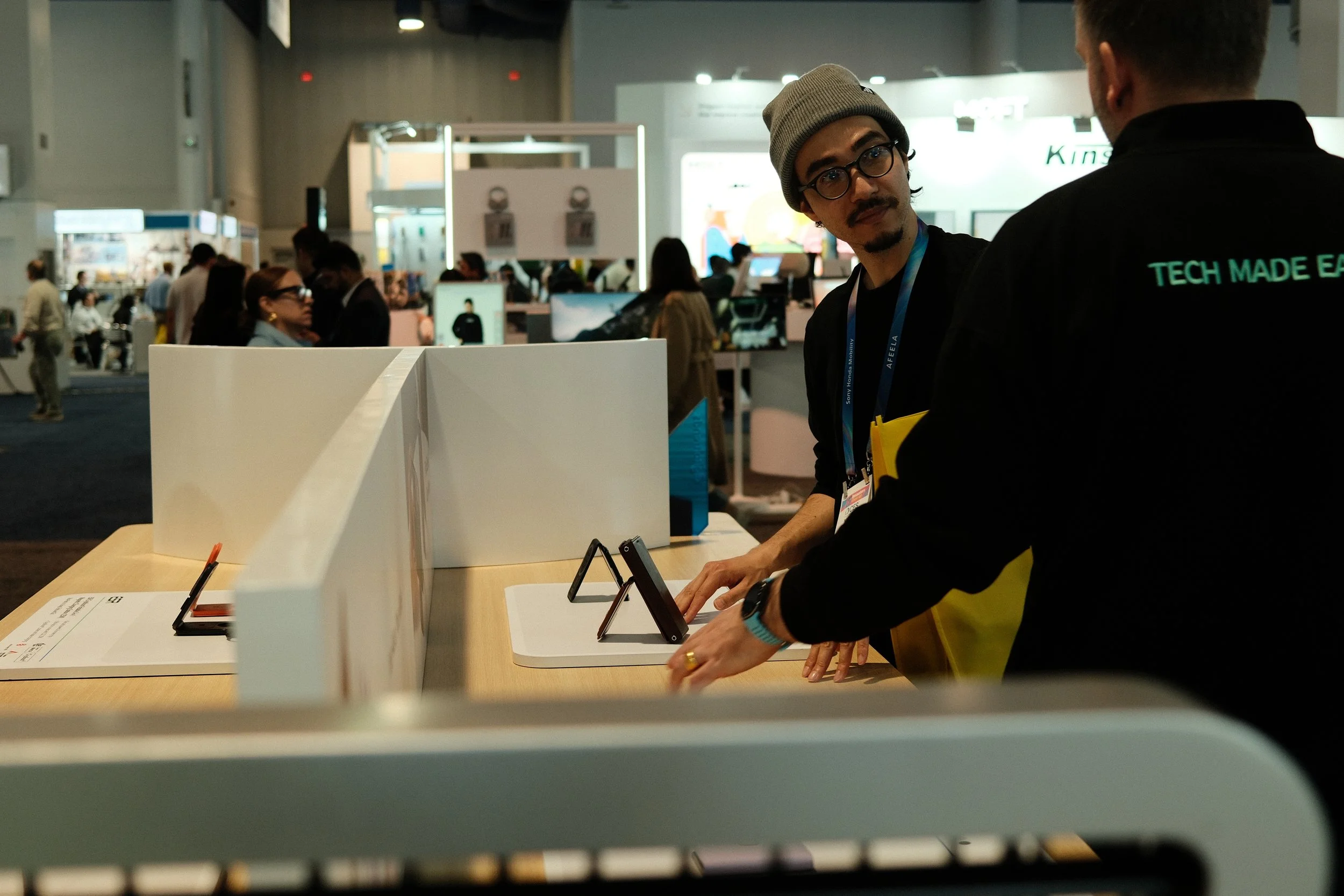Two men are at a booth displaying electronic devices, with one man wearing glasses, a beanie, and a lanyard, looking at the camera, and the other man facing away, wearing a black shirt with green text that reads 'TECH MADE EASY'.
