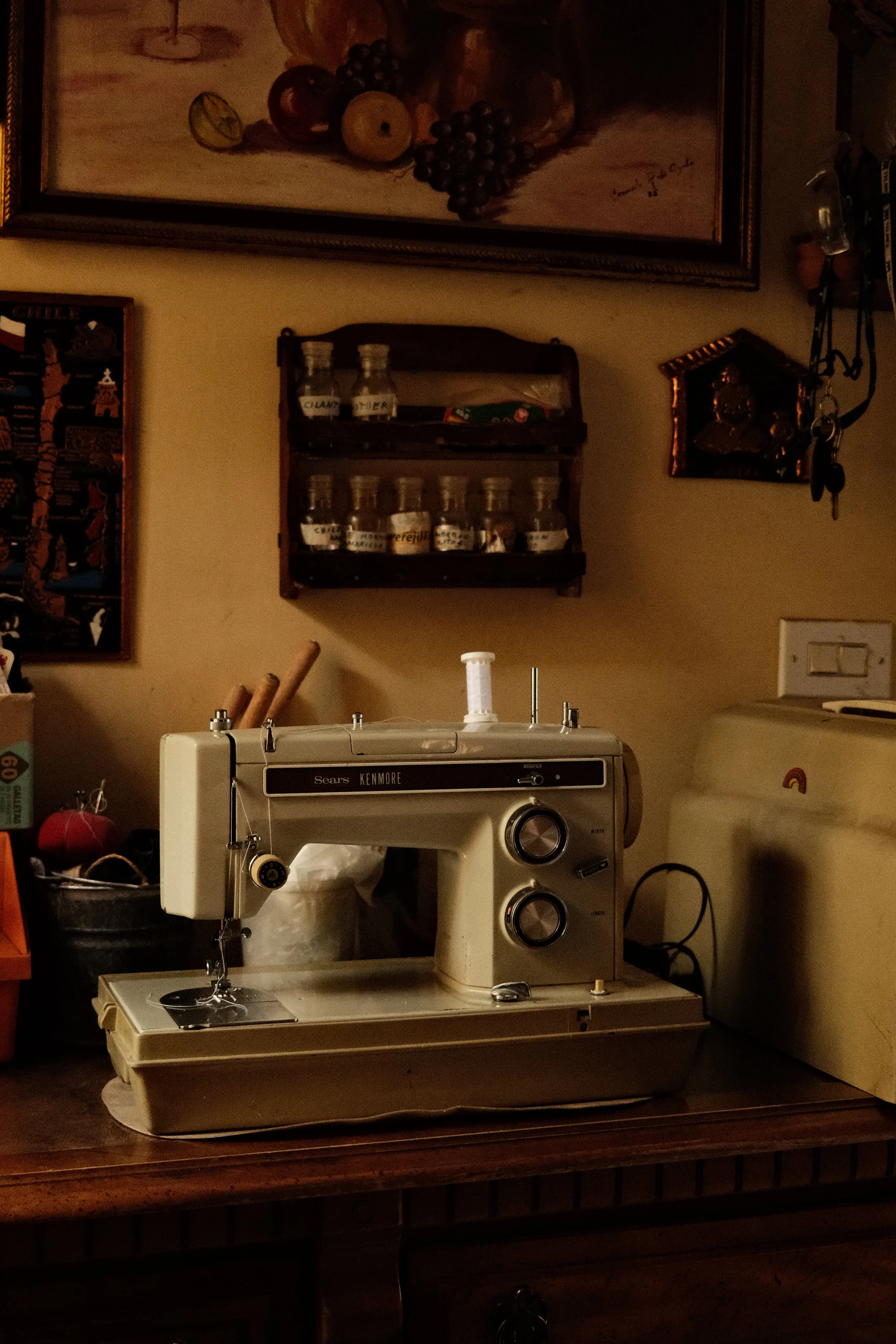A vintage sewing machine on a wooden table in a room with wall decorations and a small wall shelf holding jars.