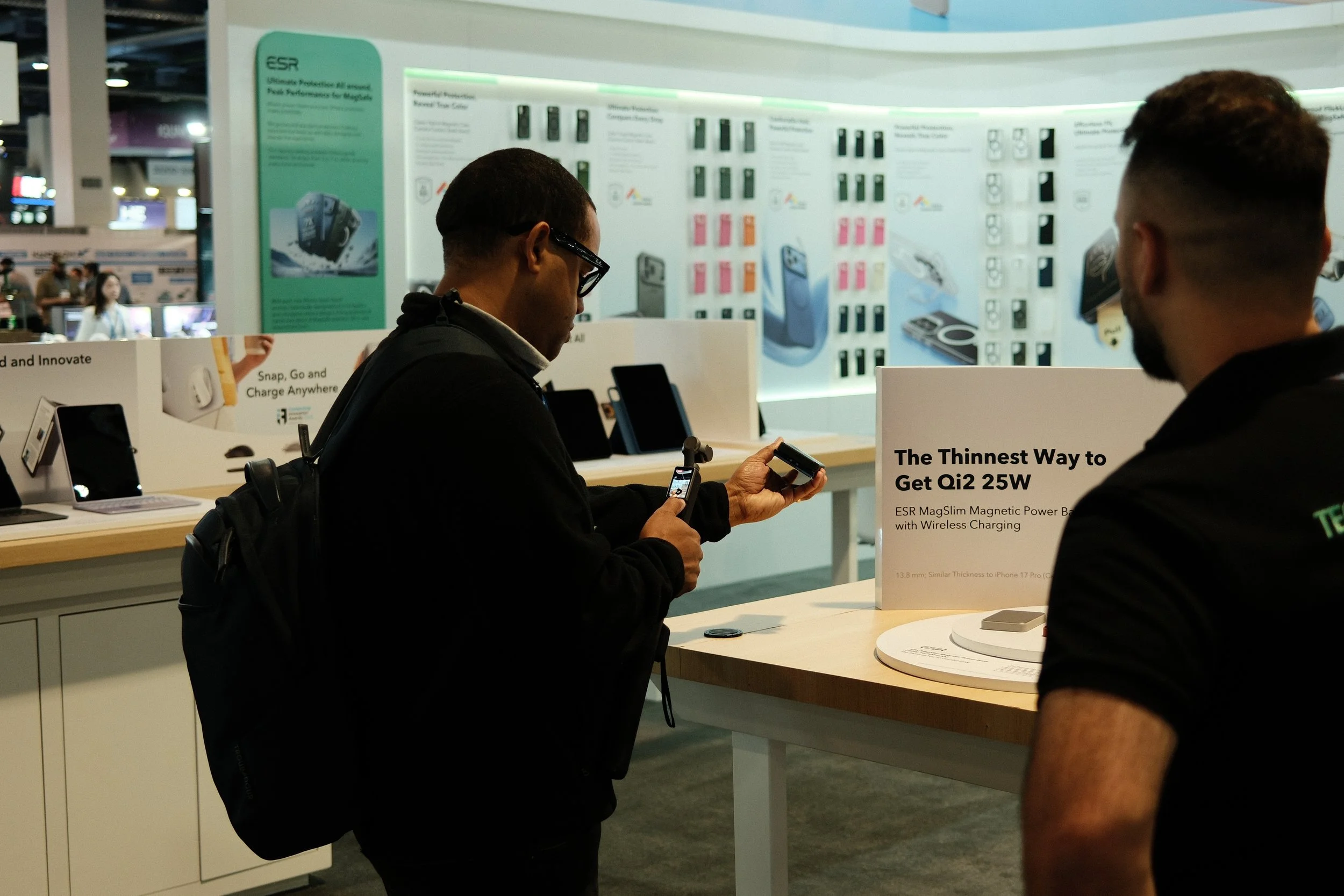 Two men at an electronics booth, one examining a product and the other observing, with display posters and products in the background.