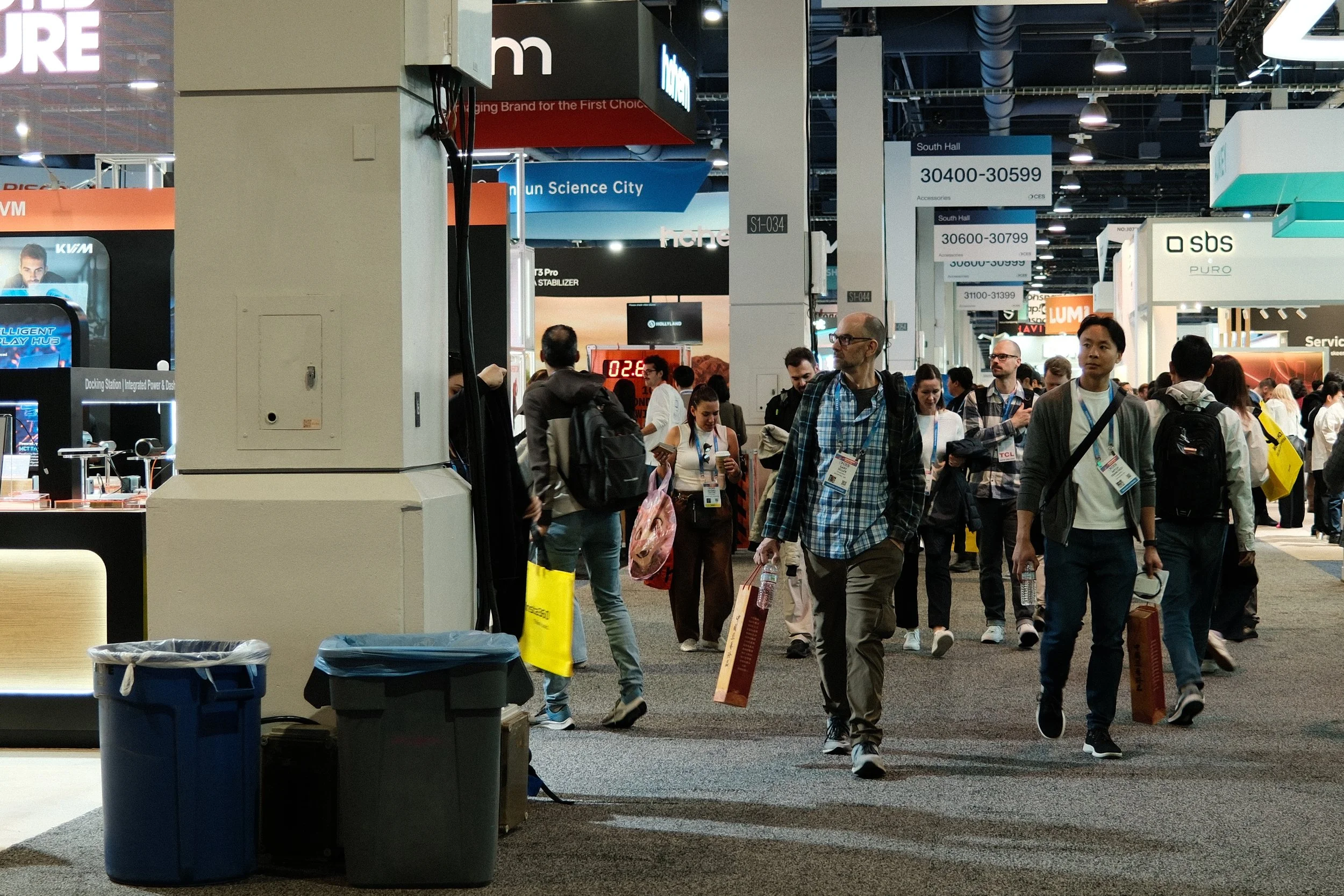 A busy convention center with people walking and carrying bags. Various exhibition booths and signs are visible, including signs labeled 'South Hall' and booth numbers. Some attendees are looking at their phones as they walk.