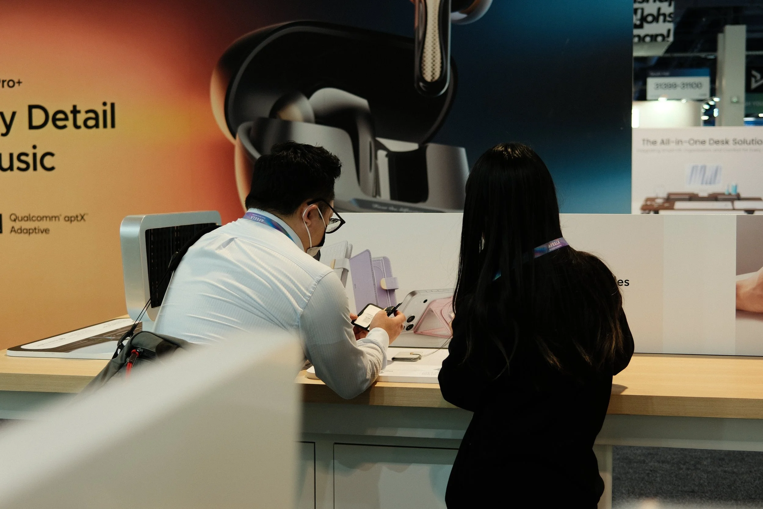 Two people at a store counter, one man wearing a white shirt and face mask, and a woman with long dark hair, both viewing electronic devices.