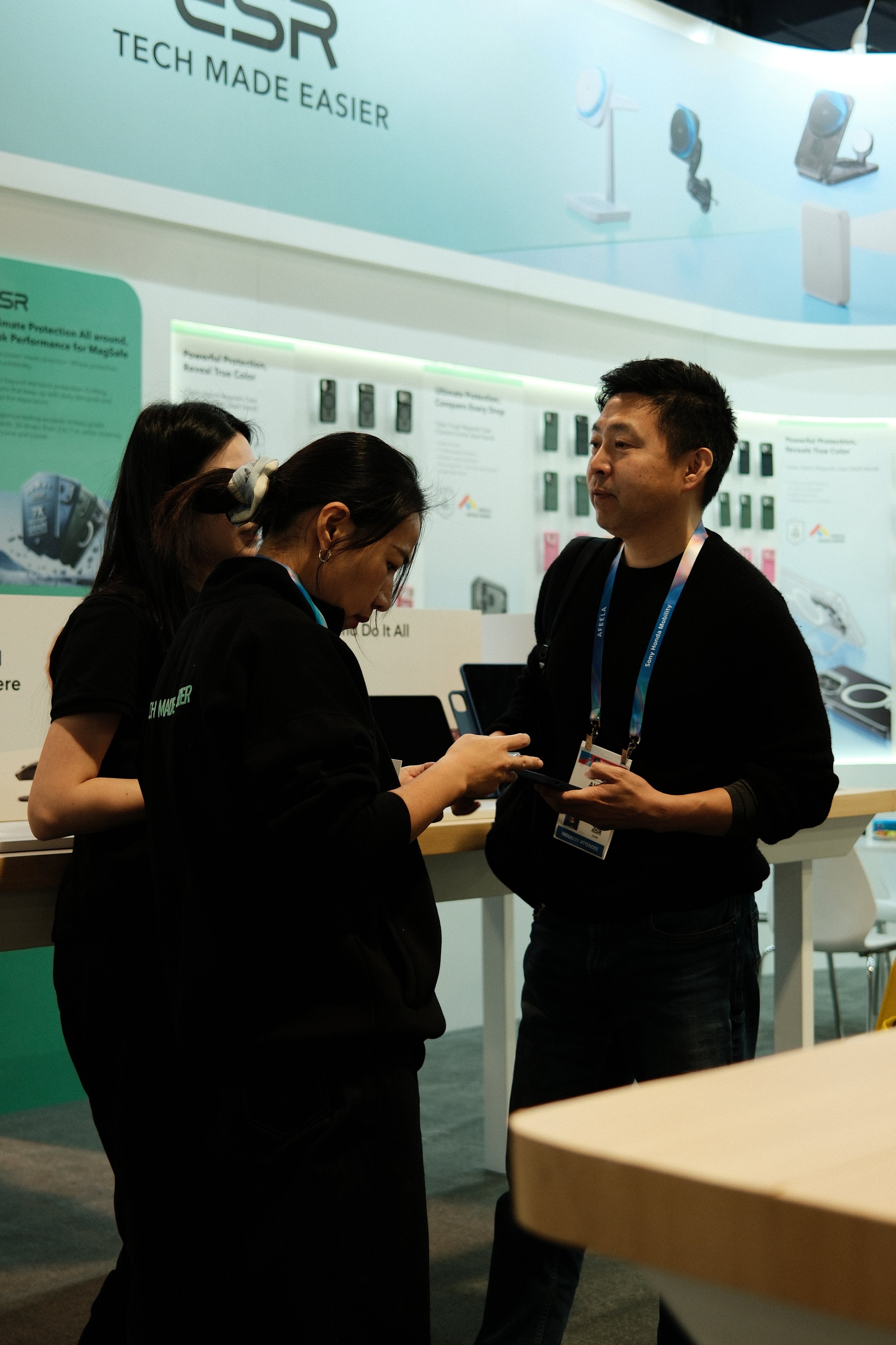 People at a tech trade show booth discussing products with a large display showing cameras and electronic devices in the background.