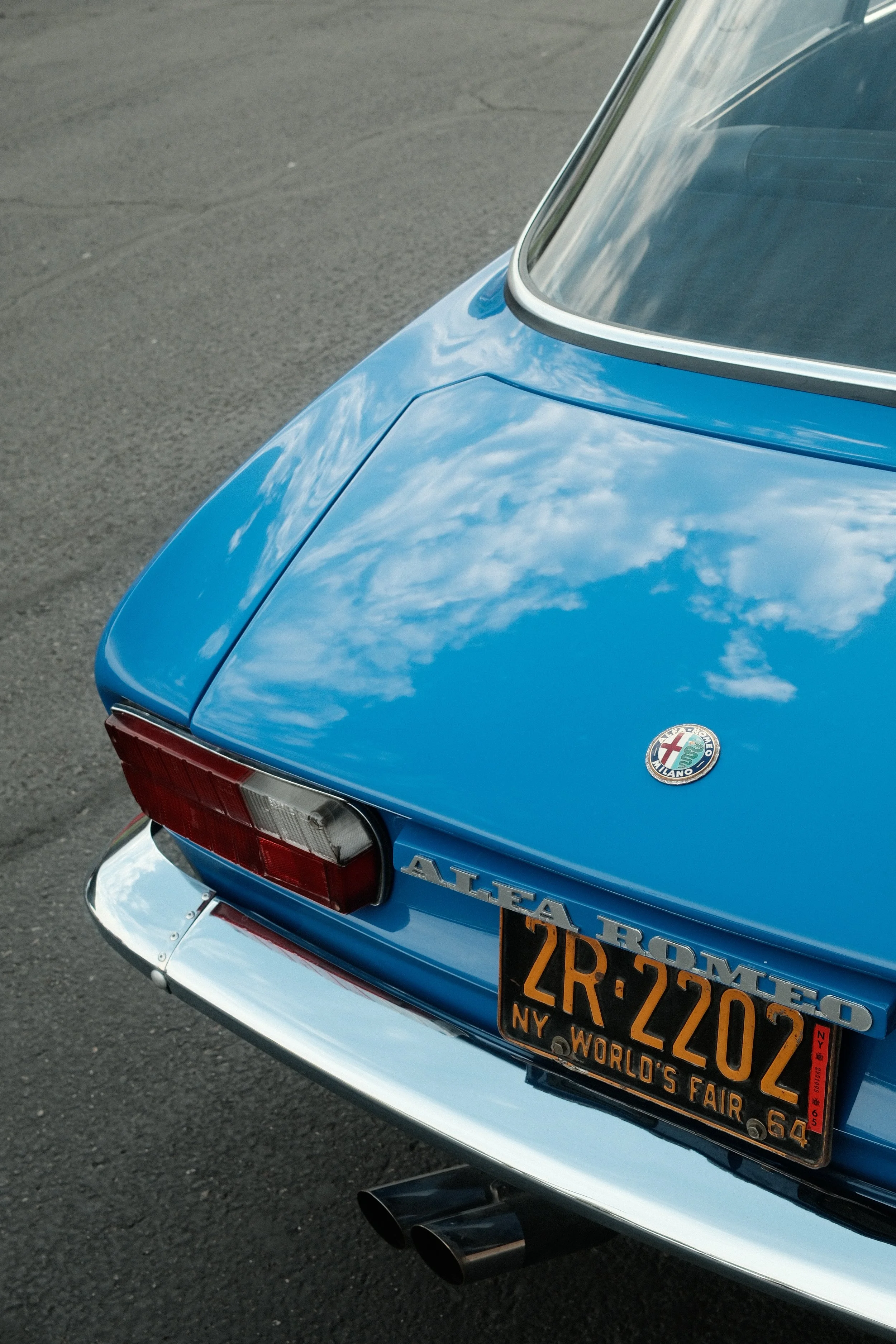 Rear view of a vintage blue Alfa Romeo car with a New York license plate that reads 2R-2202, parked on an asphalt surface, reflecting the sky and clouds on its shiny surface.