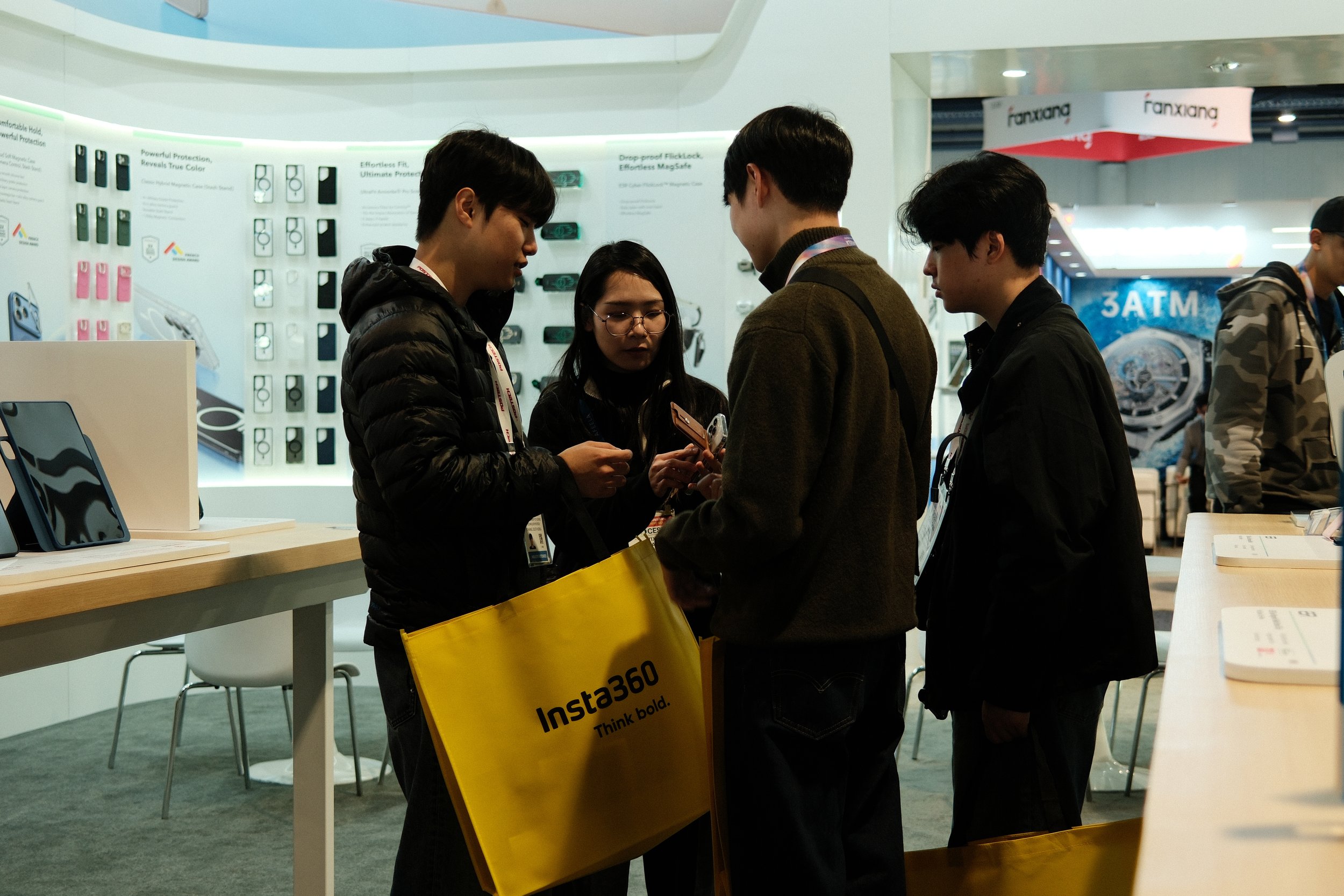 Group of four people standing at a booth, looking at their phones. They are inside an electronics or tech Expo, with displays of phone cases and devices in the background. One person is holding a yellow tote bag with the text 'Insta360 Think bold.'