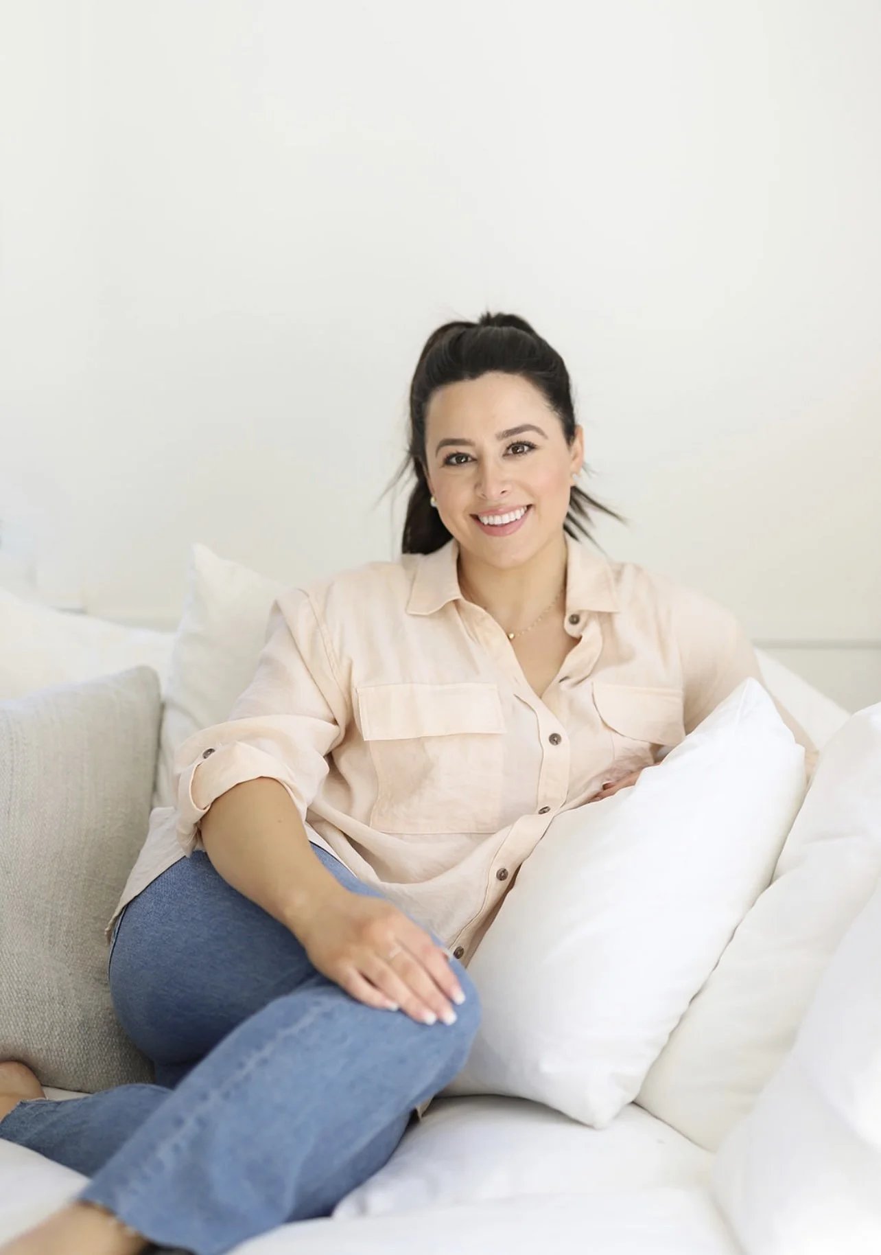 A woman with dark hair in a ponytail, smiling, sitting comfortably on a white couch with white pillows, wearing a light-colored button-up shirt and blue jeans.