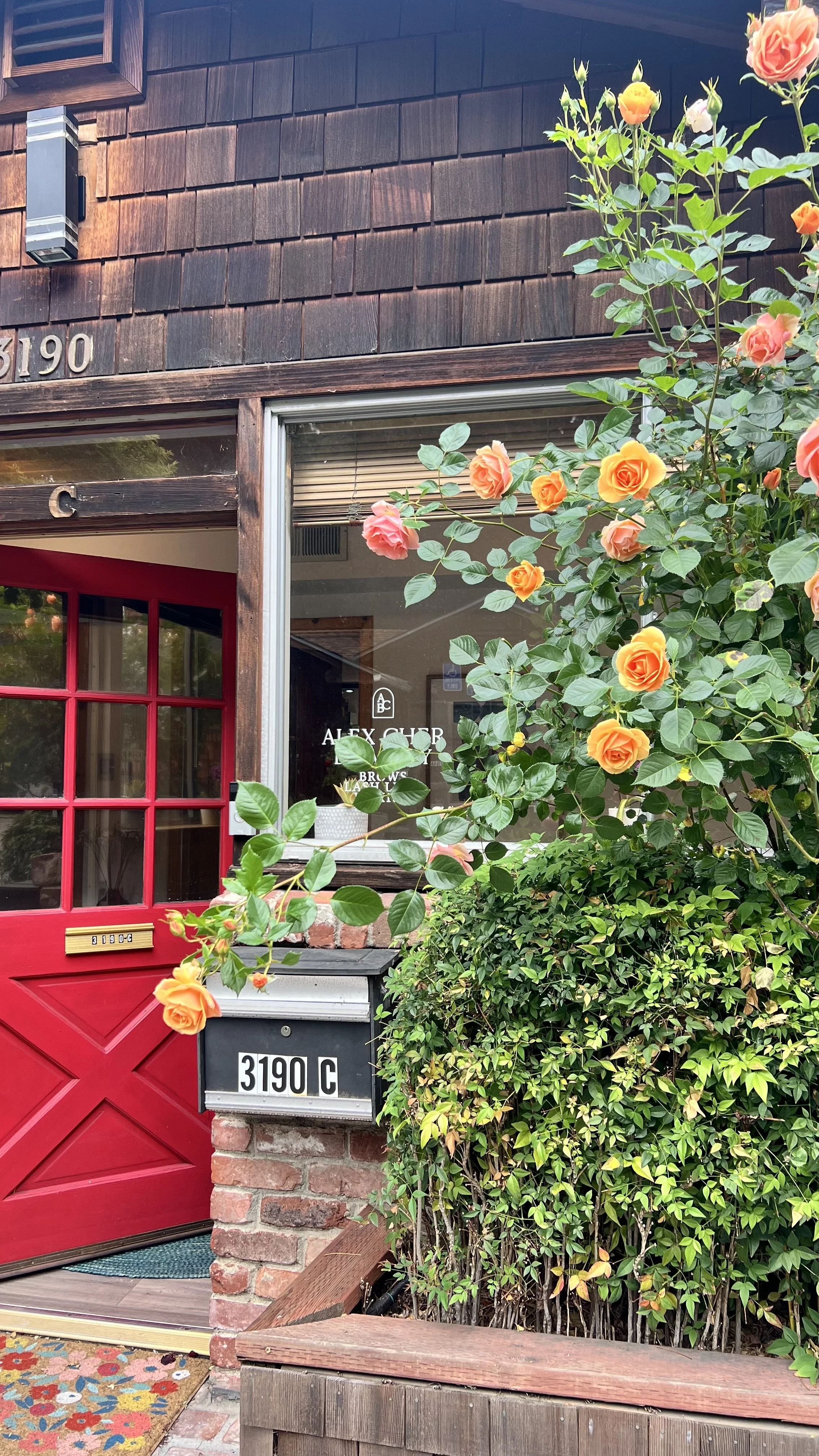 The image shows the exterior of a building with a dark brown wooden facade and the house number 3190C. There is a bright red front door with a mailbox attached below the house number. To the right of the door, there is a large window with the words 'ALEX CLUB' and some smaller text inside. A lush green bush with orange and pink roses partially obscures the lower right part of the house. A decorative multicolored mat is placed in front of the door.