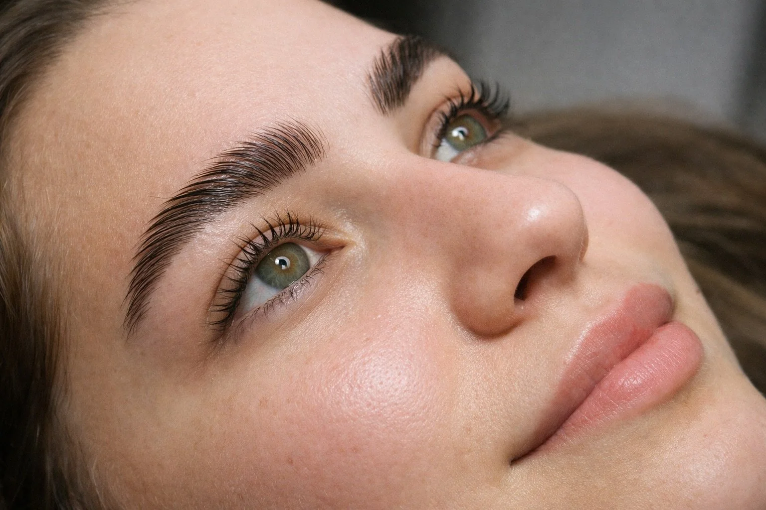 Close-up of a woman's face, highlighting her well-groomed eyebrows, long eyelashes, and clear skin, with a focus on her eyes and lips.