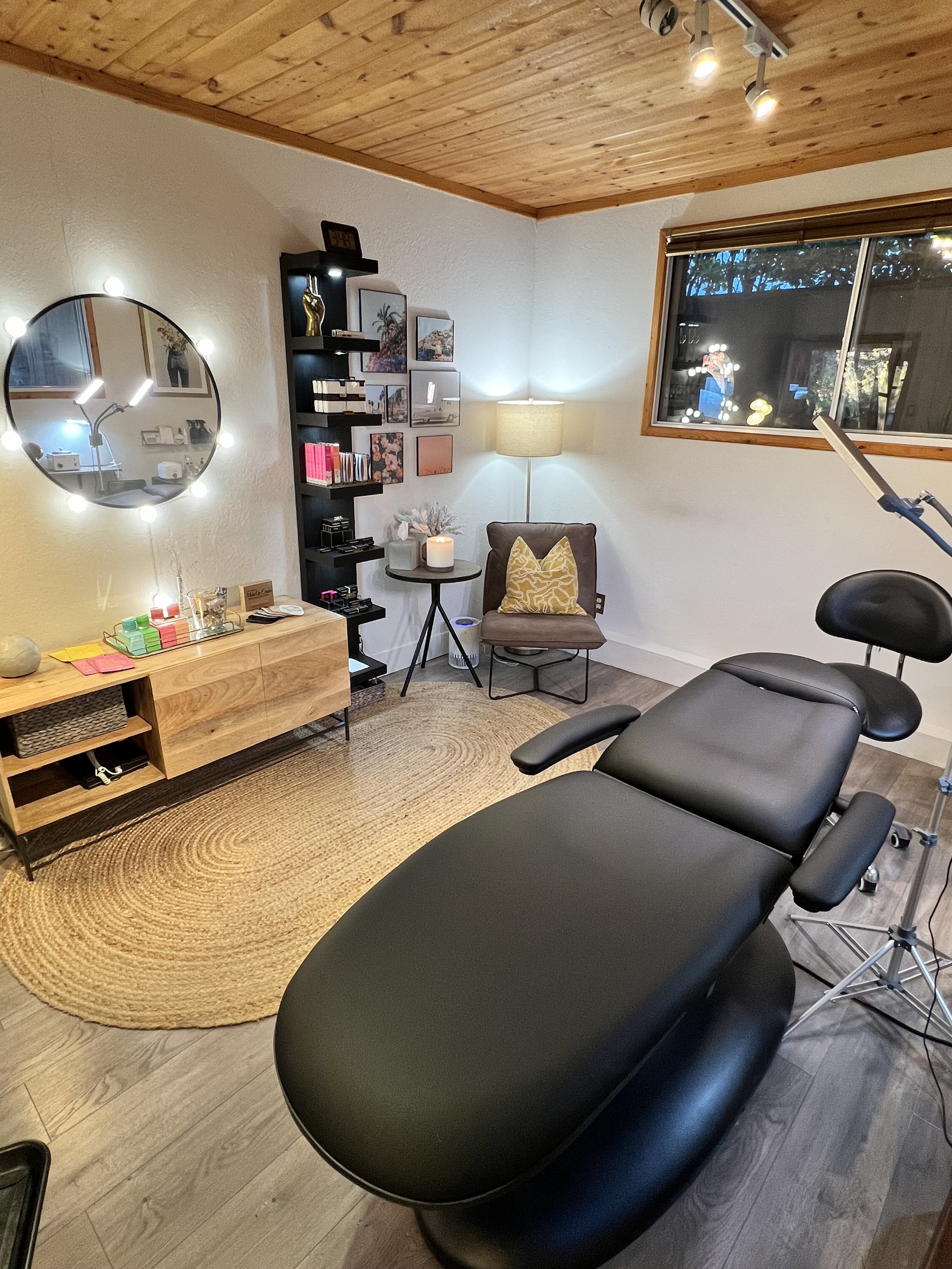 A therapy or massage room with a black reclining chair, a wooden sideboard, a round mirror with lights, a shelving unit with books and decor, a large window, a floor lamp, and a decorative rug.