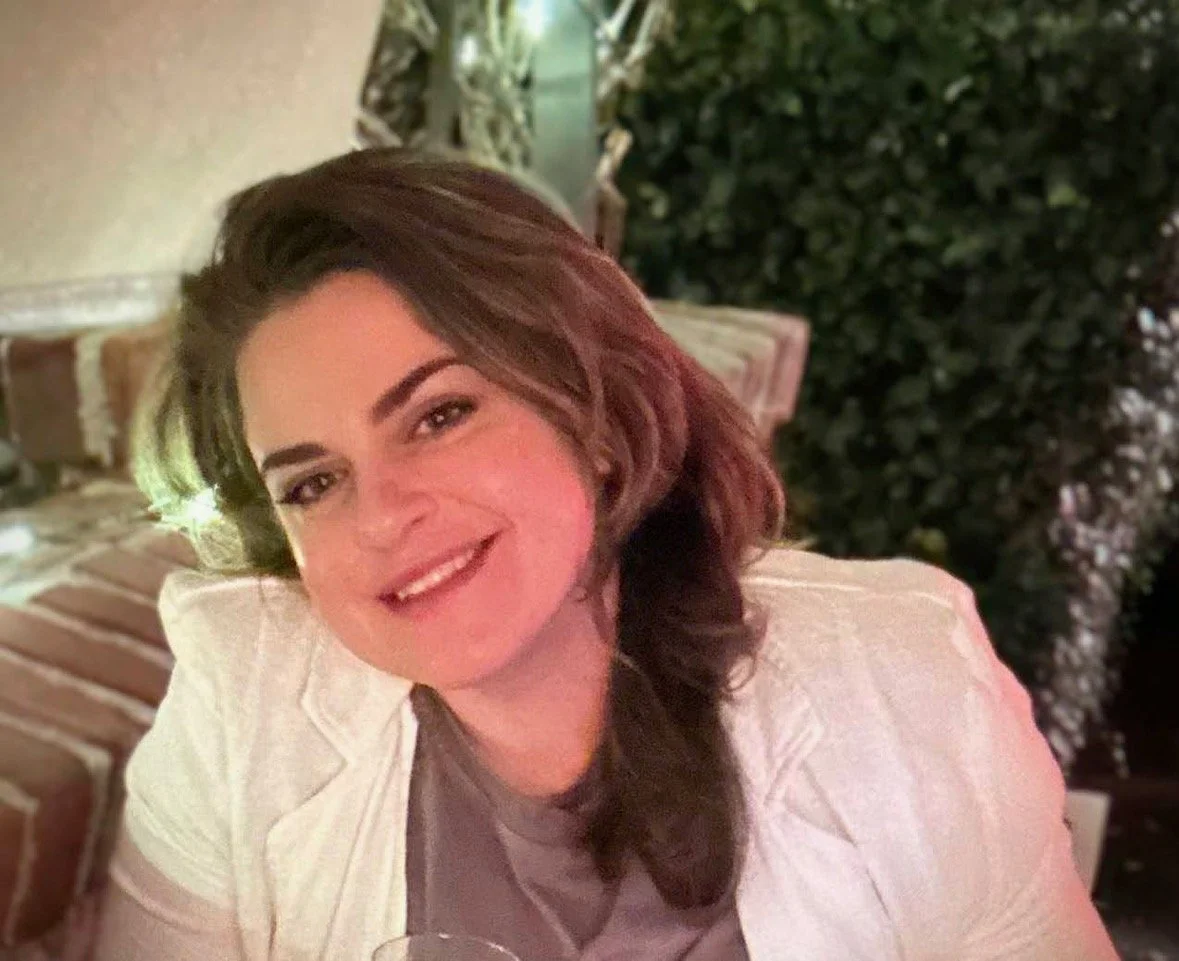 A woman with shoulder-length brown hair and a white blazer smiling at the camera, sitting outdoors near a brick wall and green foliage.