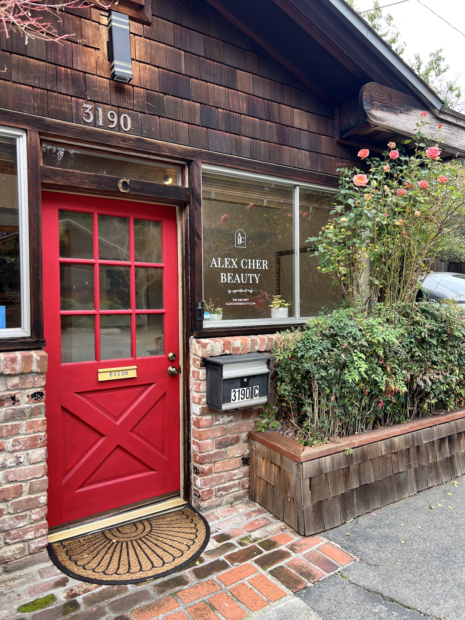 Red door with glass panes at the entrance of Alex Cher Beauty salon, located at 3190 C. The exterior features a brick and wood facade, a black mailbox in front, and pink roses bush to the right.