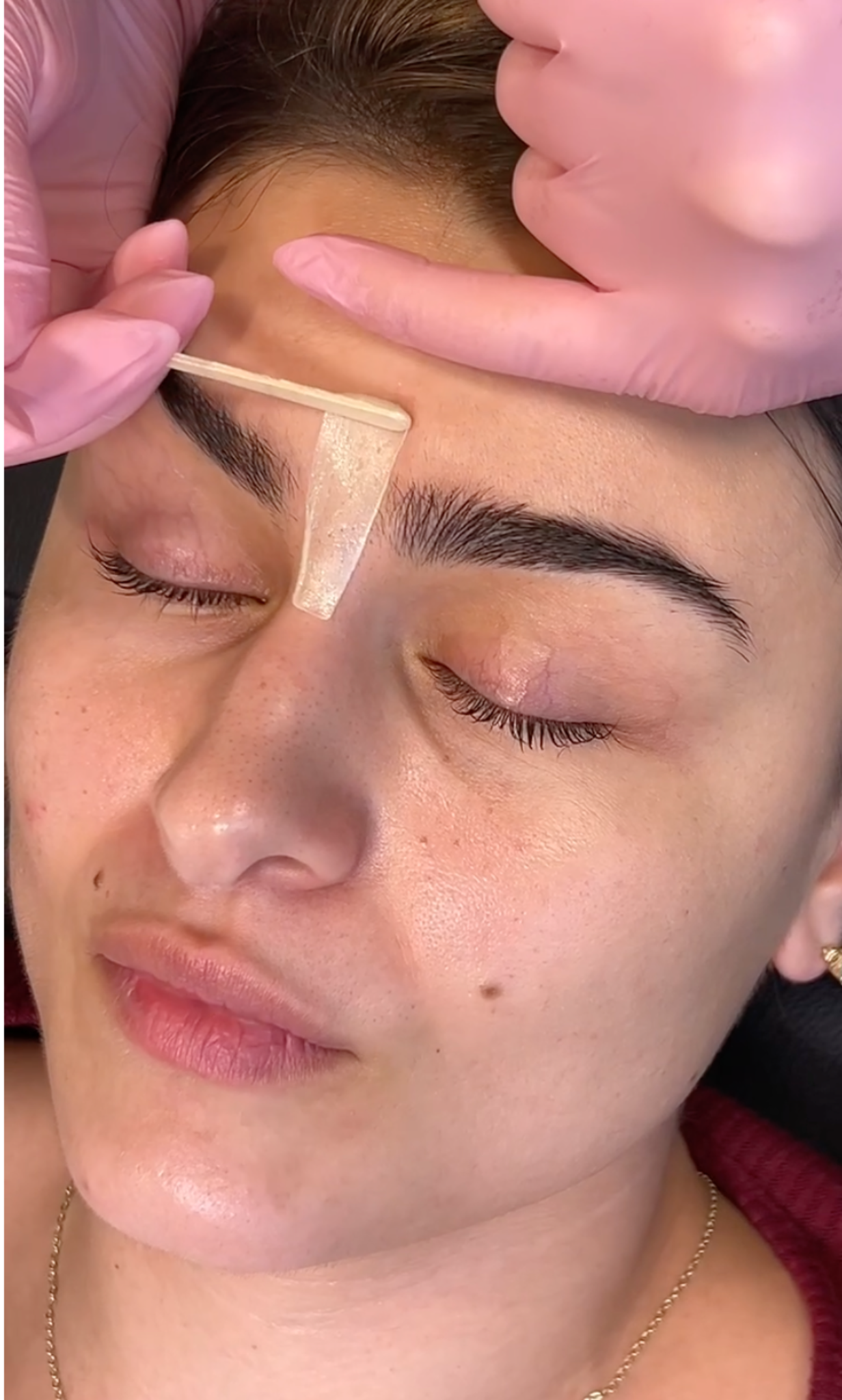 A woman receiving a waxing treatment on her eyebrow, with a gloved technician applying wax with a spatula on her eyebrow.