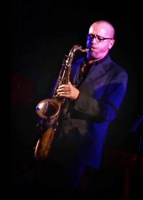 A man with glasses in a dark suit playing a saxophone on stage with low lighting.
