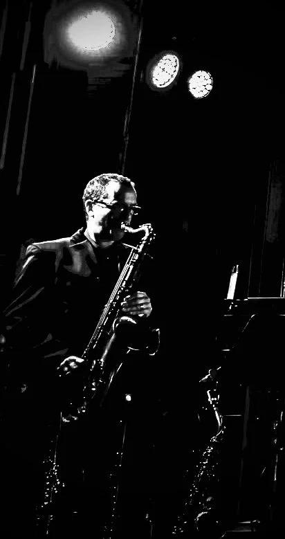 A man in a suit playing a saxophone on a dimly lit stage, with two clocks visible on the wall behind him.