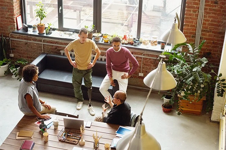 An overhead view of a small group of colleagues having an informal discussion around a table with laptops in a casual, plant-filled office space.