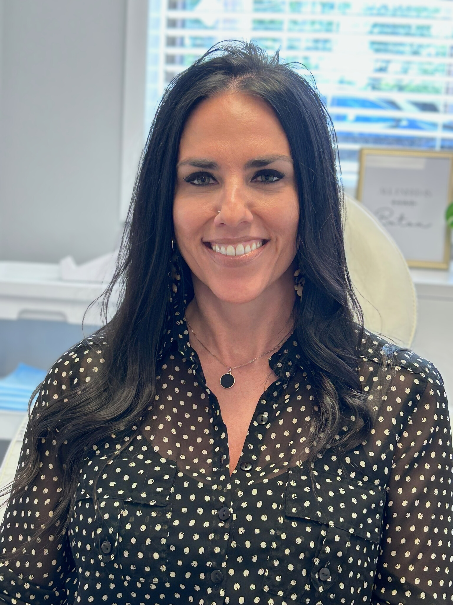 A woman with long dark hair, wearing a black polka dot blouse and a necklace, smiling in an office setting.