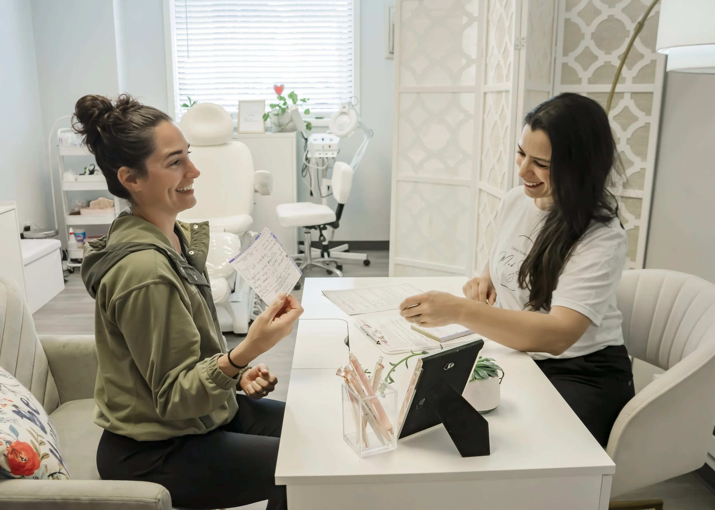 A woman at a medical office presenting a handwritten note to another woman sitting across the desk, both smiling.