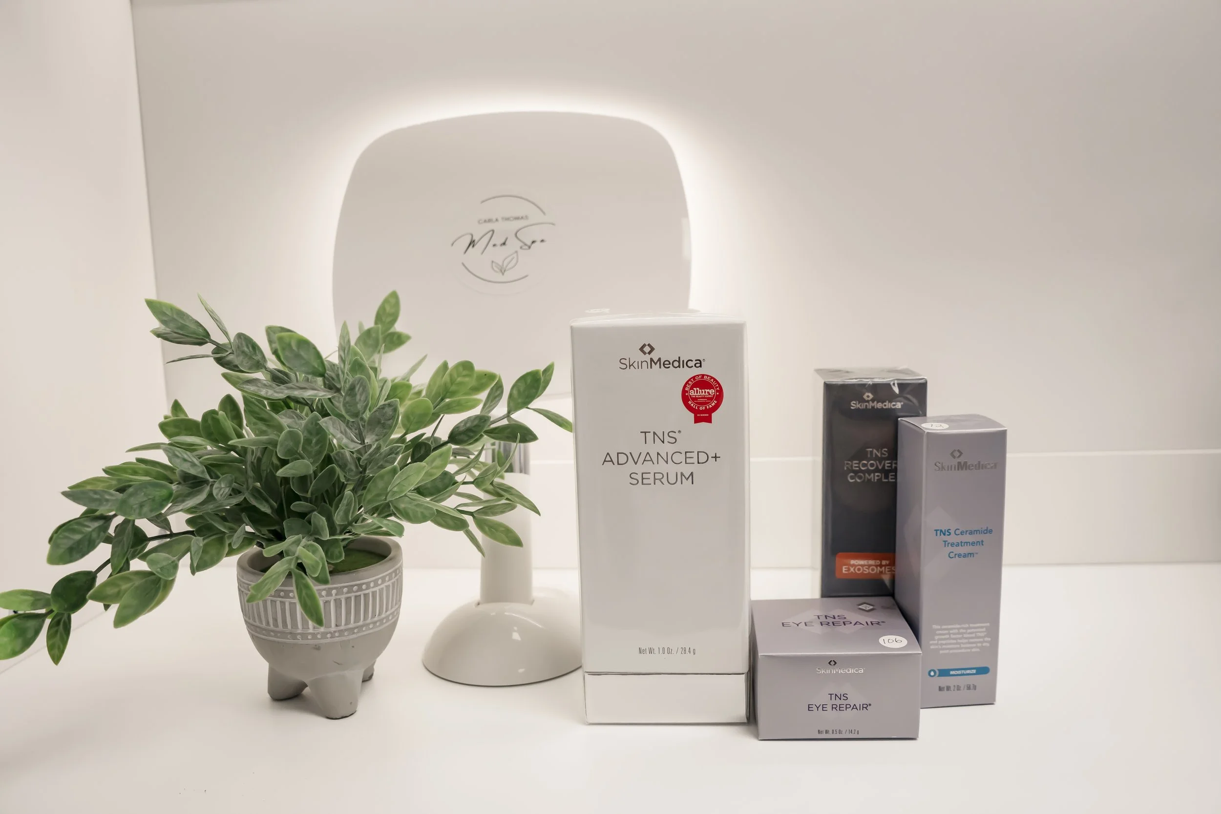 Skincare products including SkinMedica TNS Advanced+ Serum, TNS Recovery Complex, TNS Eye Repair, and TNS Ceramide Treatment Cream arranged on a white surface with a potted plant and a mirror in the background.