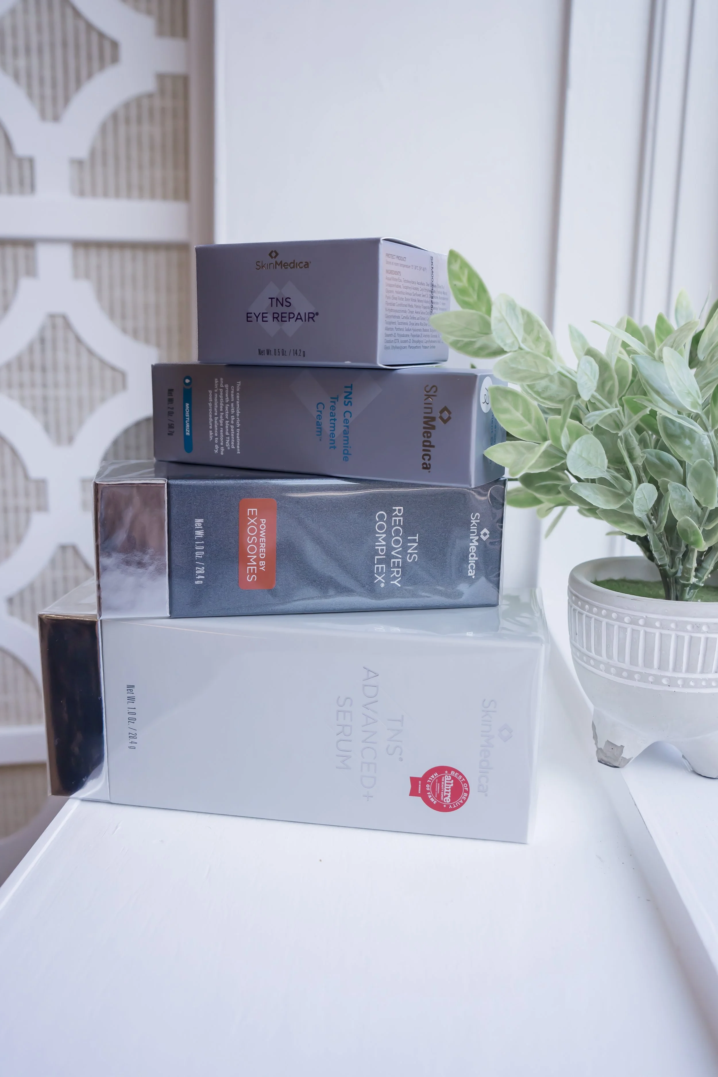 A stack of SkinMedica skincare products including TNS Advanced+ Serum, TNS Recovery Complex, Ceramide Treatment Cream, and Eye Repair, styled next to a decorative potted plant on a white counter.