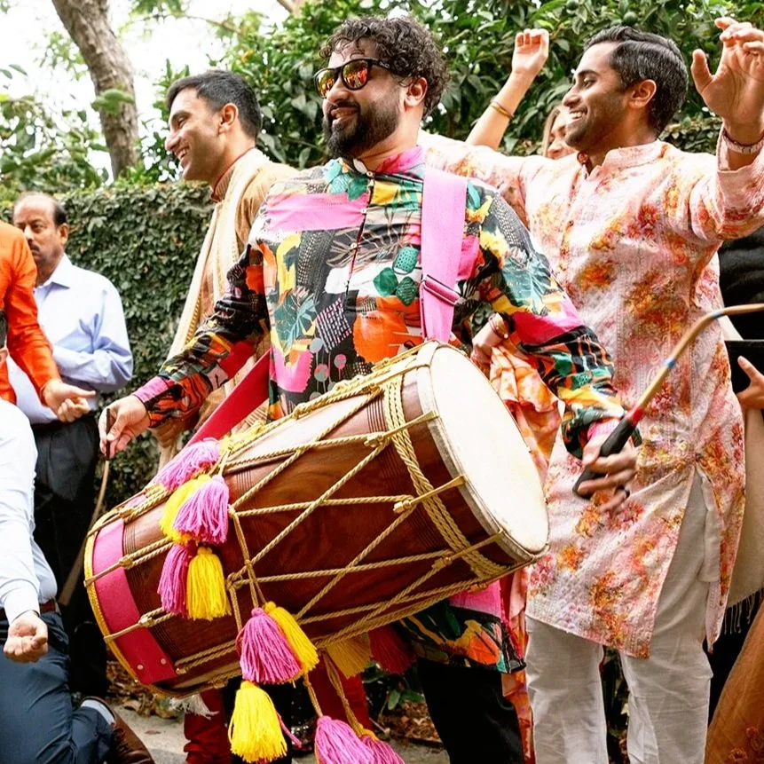 A group of men dancing outdoors at a Baraat with Vin Rocc playing a Dhol, wearing a colorful kurta and enjoying the celebration or grooms processional.