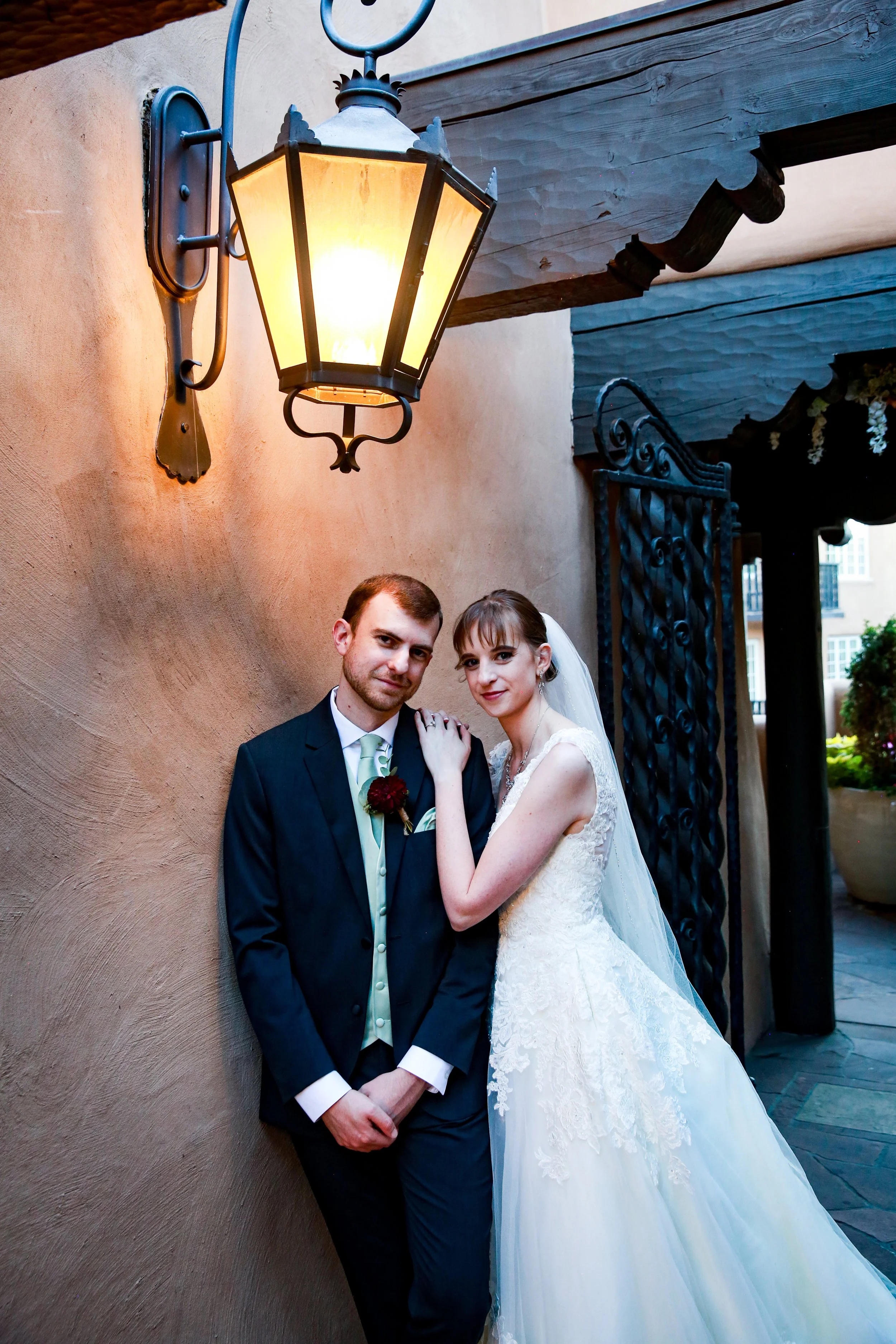 At La Fonda de Santa Fe, the top wedding venue in all of Santa Fe, a bride and groom pose outside in the terrace with stunning views of the Cathedral of Saint Francis, illuminated by a warm light fixture overhead during dusk.