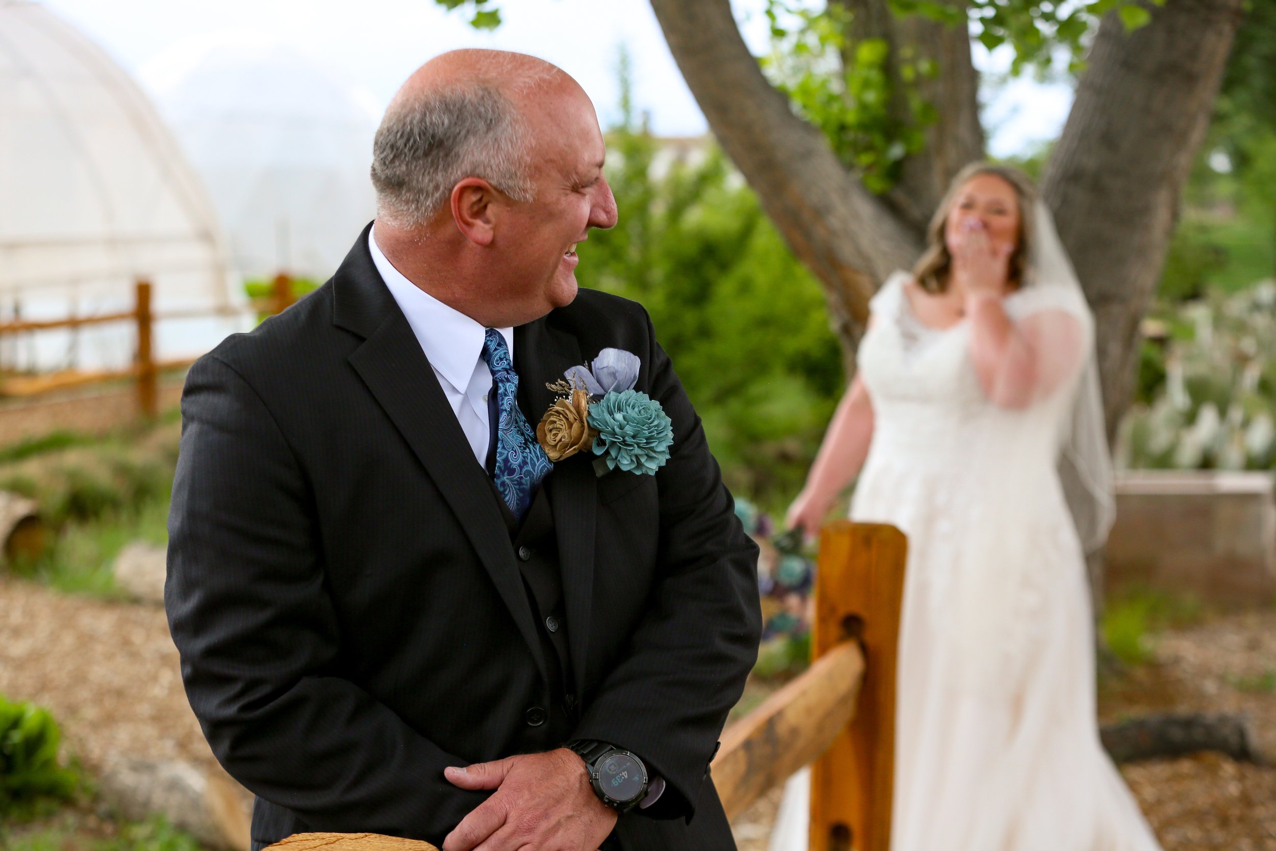 A man in a black suit with a teal boutonniere looking to the side, smiling, during an outdoor wedding ceremony. A woman in a white dress stands in the background near trees.