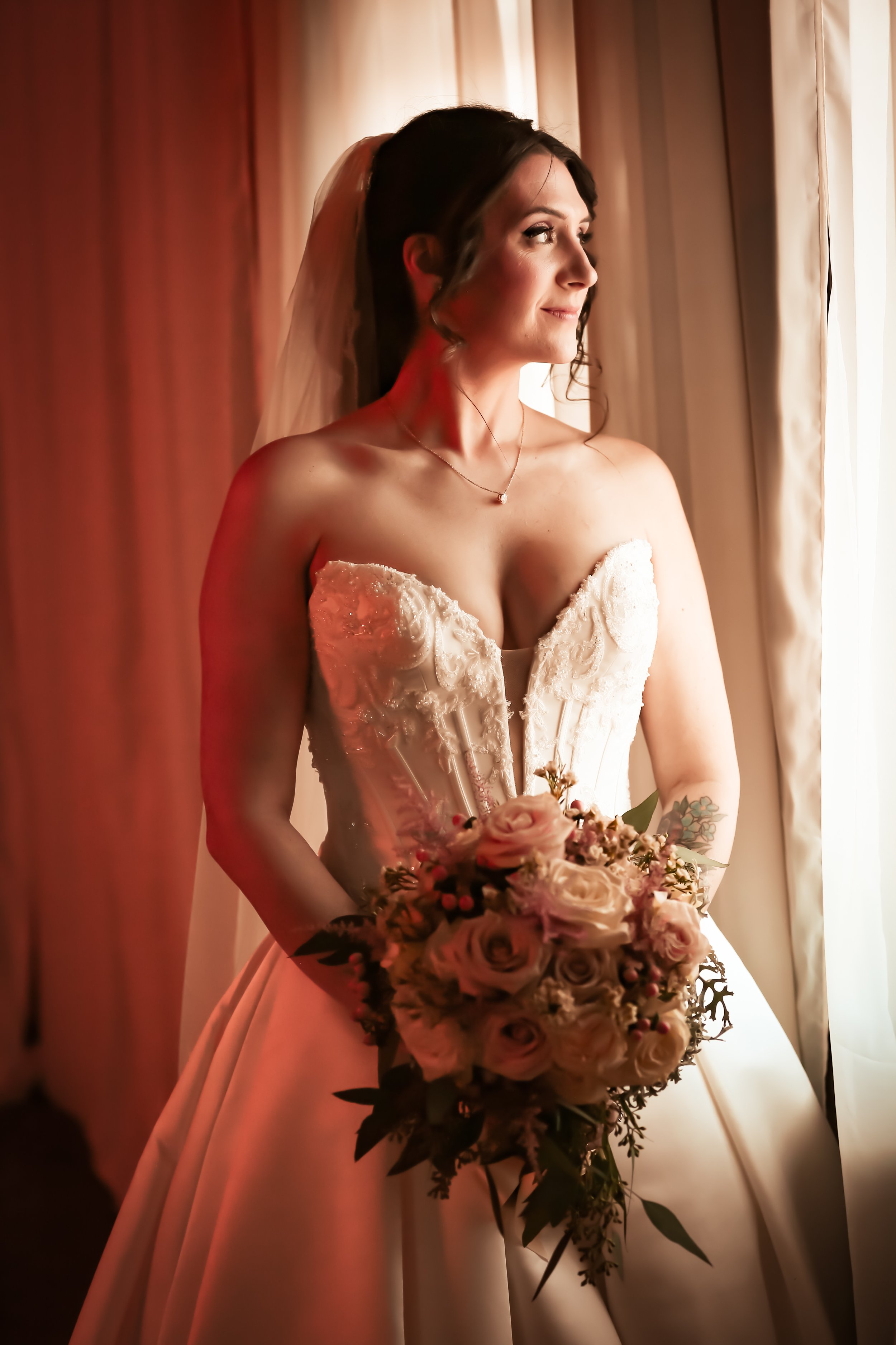 A bride in a strapless wedding gown holding a bouquet of pink and white roses, standing by a window at the river ranch in williston nd