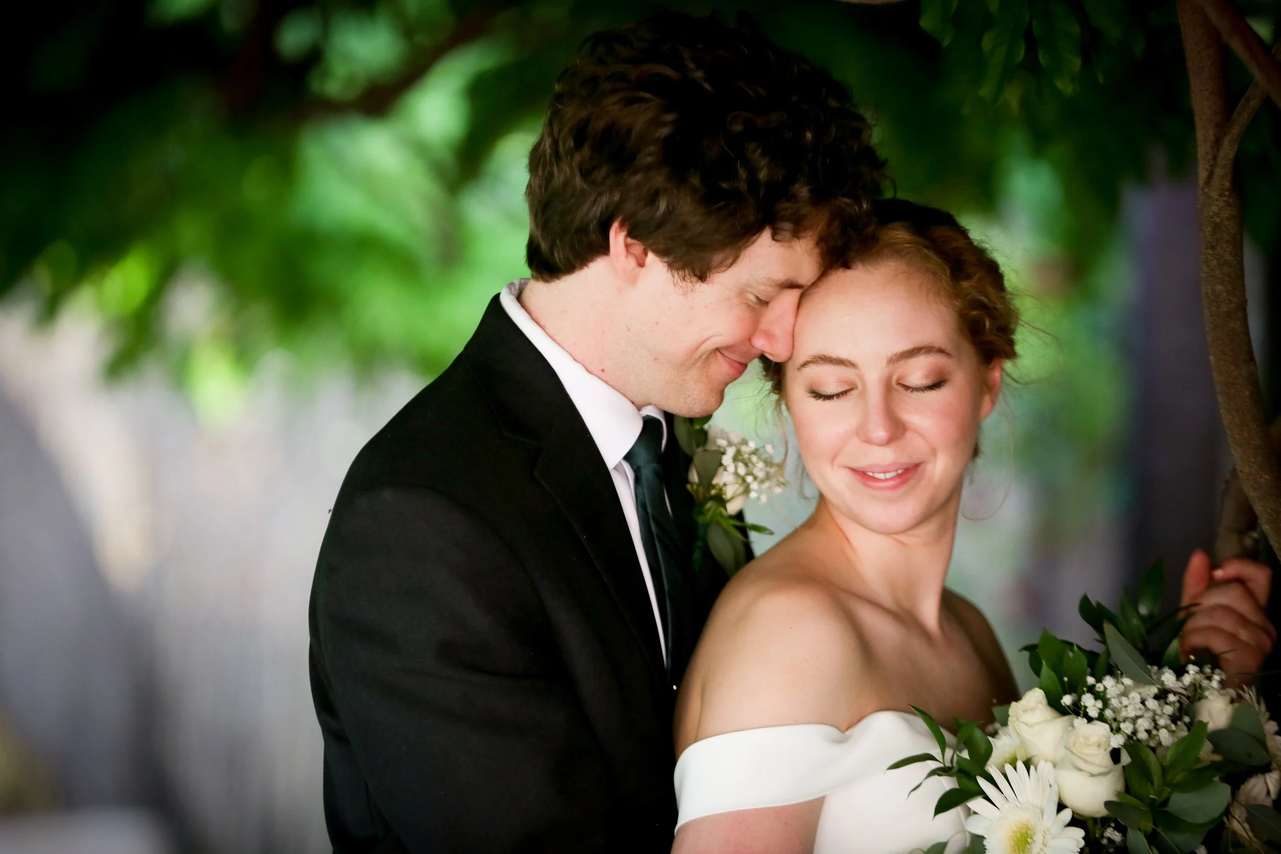 Los Poblanos - A bride and groom sharing a tender moment with their foreheads touching in a lush outdoor setting, the bride holding a bouquet of white and green flowers.