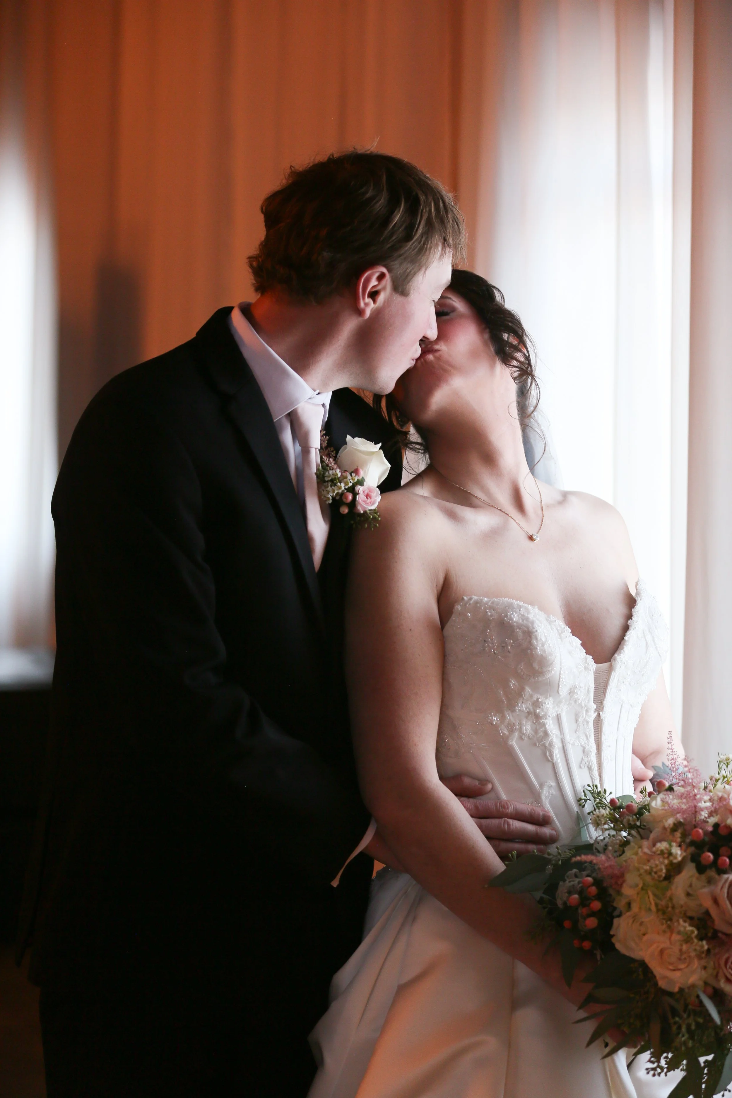 Bride and groom sharing a kiss beside a window during their wedding portrait session