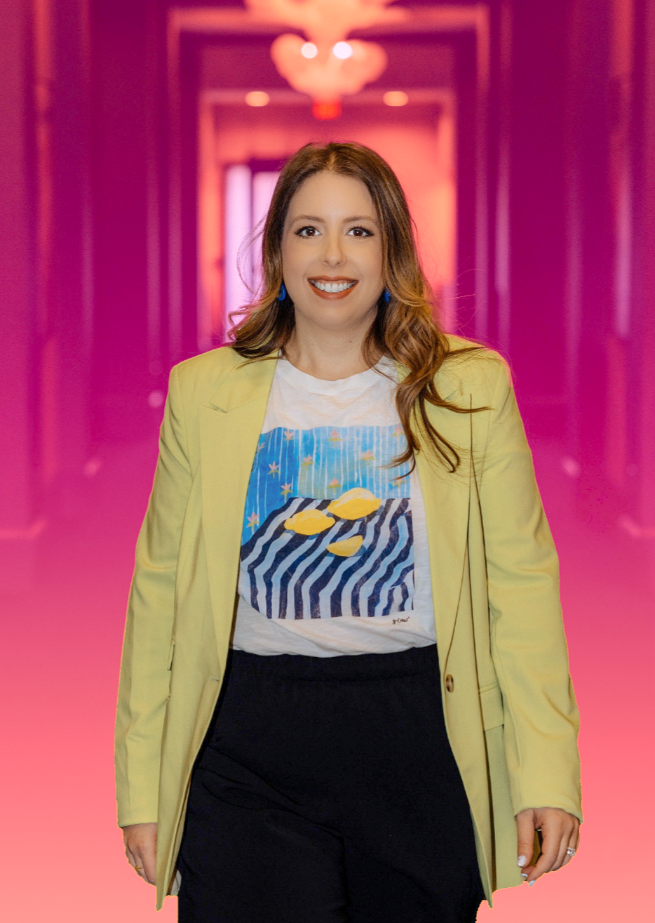A woman with light skin, long wavy brown hair, wearing a yellow blazer, a white graphic t-shirt with a colorful design, and black pants, smiling at the camera. The background is a pink-lit hallway with a chandelier.