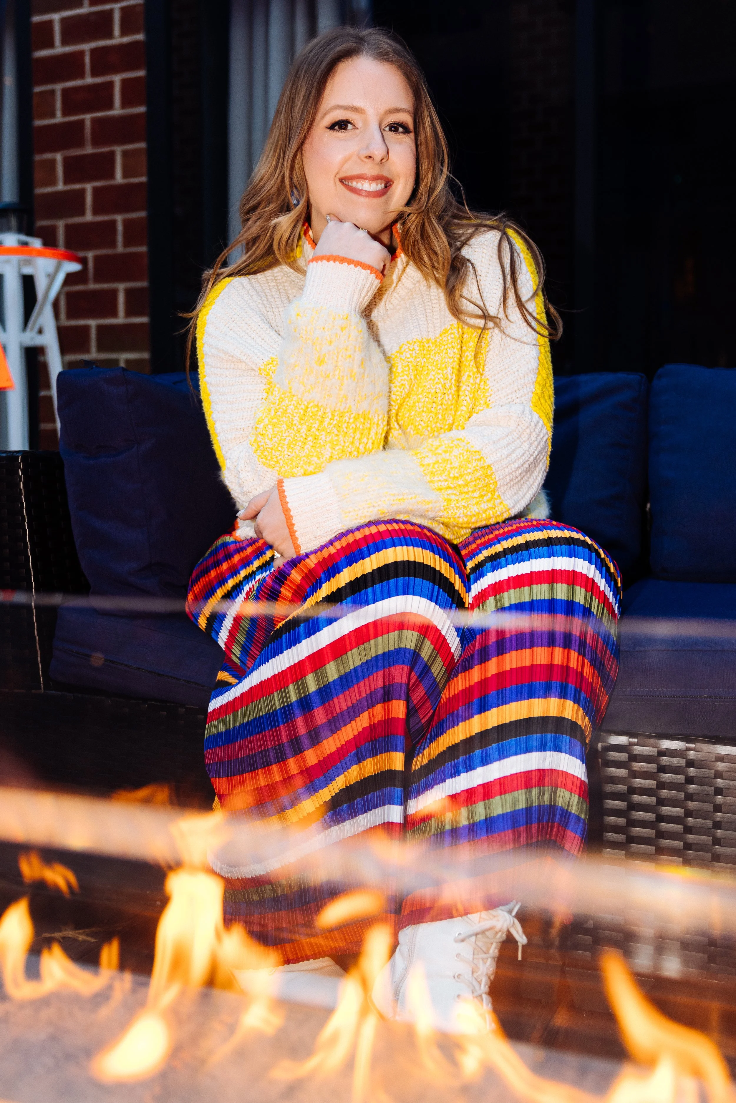 A woman with light brown hair and light skin sitting on a dark blue outdoor sofa, wearing a colorful striped pant and a yellow and white sweater, smiling with her hand resting on her chin, with a fire in front of her.