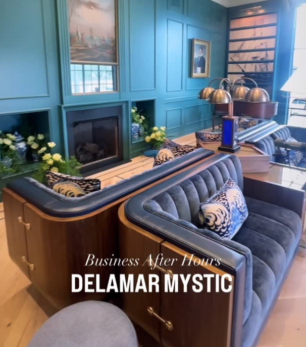 Elegant living room with teal paneled walls, a fireplace, and blue velvet tufted sofa with patterned pillows. There is a side table with gold lamps, and artwork on the walls. The image is overlaid with text reading 'Business After Hours DELAMAR MYSTIC'.