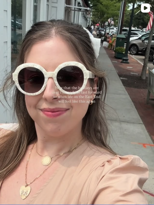 A woman taking a selfie outside on a sidewalk in Southampton New York. She is wearing large round sunglasses, a nude-colored top, and layered gold necklaces. There are parked cars, trees, and storefronts in the background.