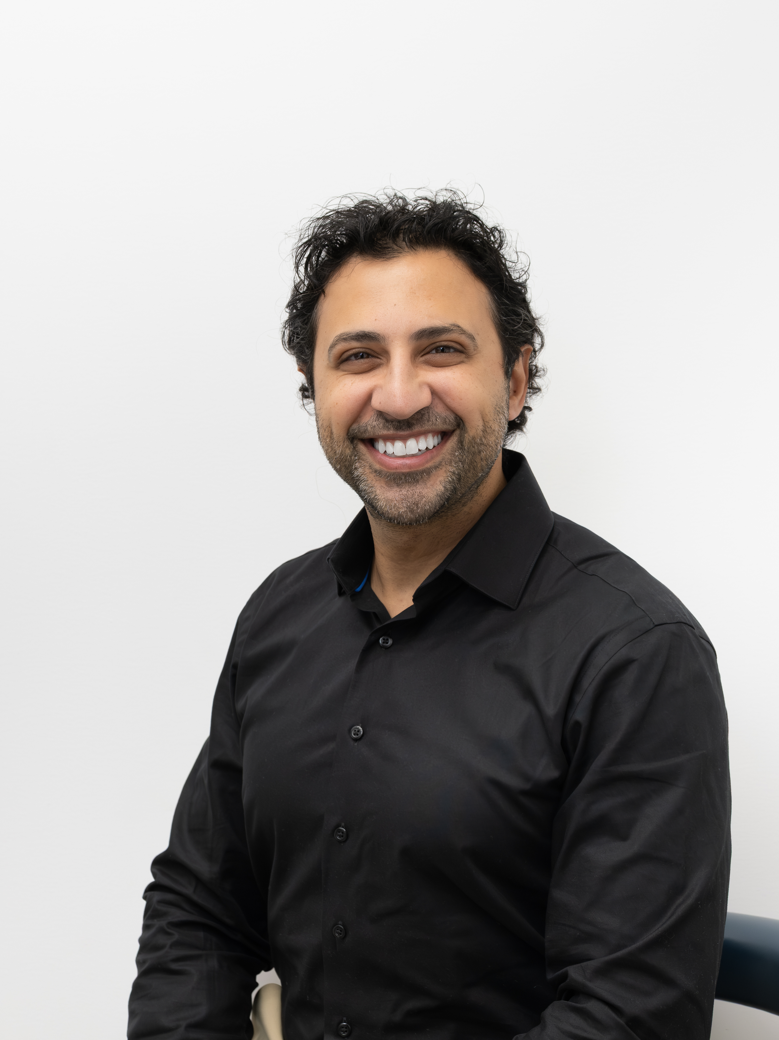 Homme souriant avec barbe et cheveux bouclés, portant une chemise noire, assis contre un mur blanc.