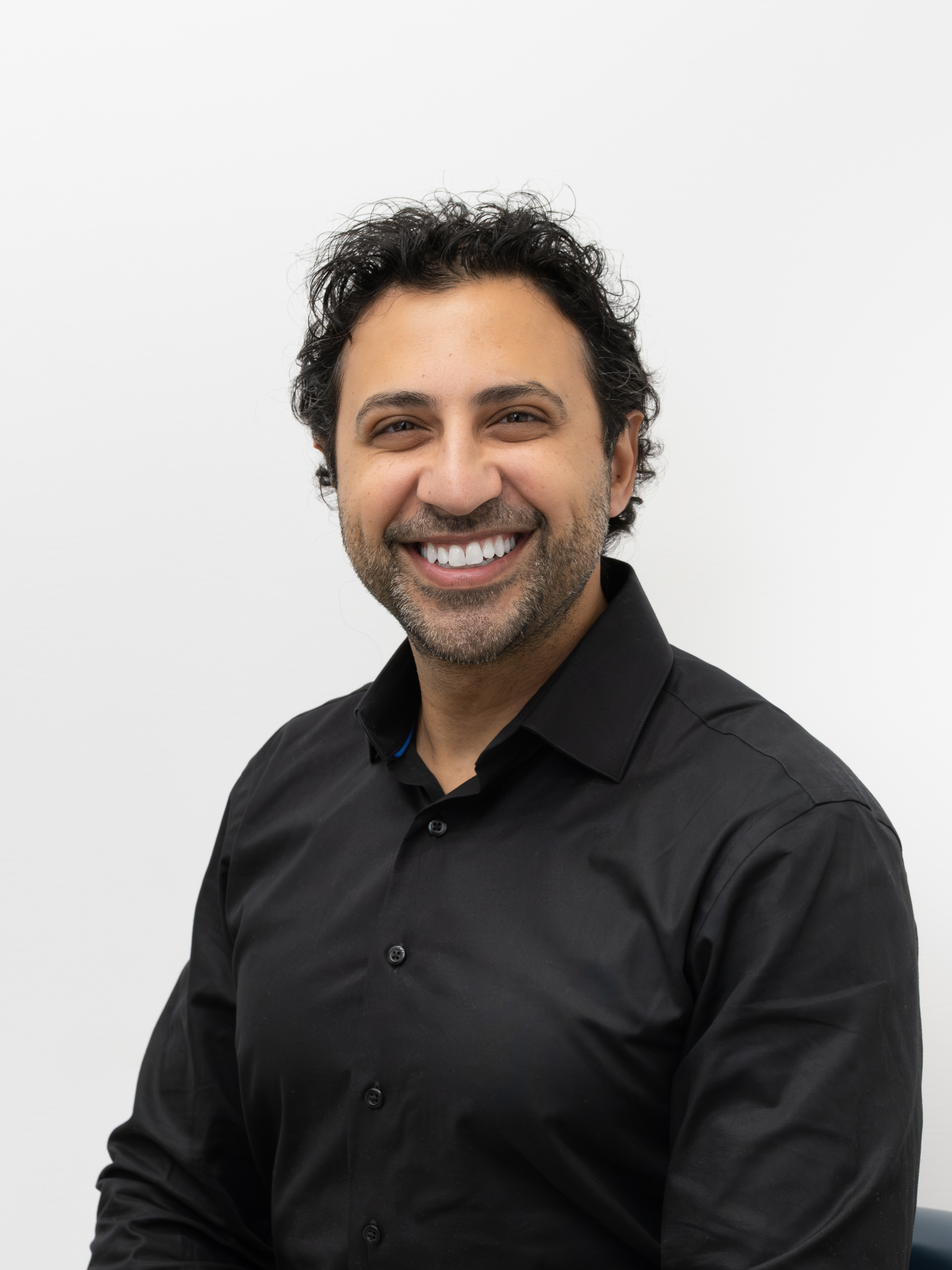 Homme souriant avec barbe, portant une chemise noire, fond blanc.