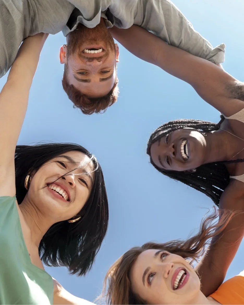 Groupe de cinq personnes souriantes qui se tiennent par la main en formant un cercle, vu du dessous contre un ciel bleu.