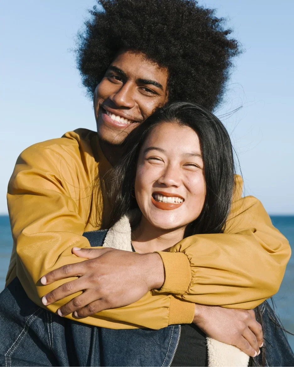 Deux personnes souriantes, un homme avec des cheveux afro et une femme aux cheveux longs noirs, s'embrassent en plein air avec la mer en arrière-plan.