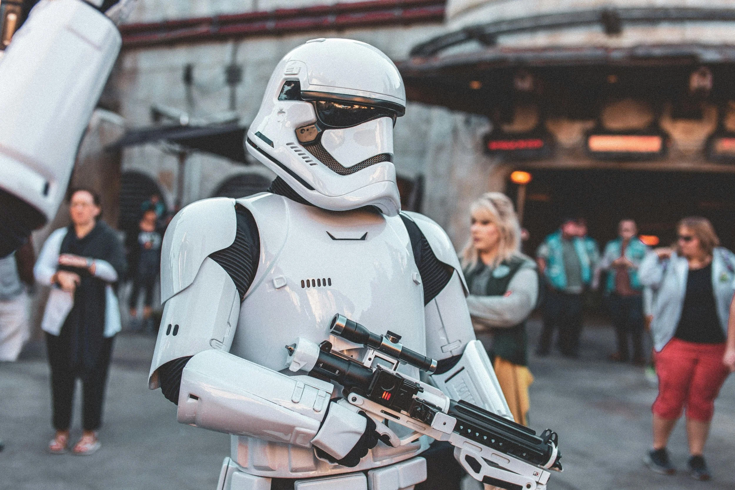 A person dressed as a Stormtrooper from Star Wars holding a futuristic weapon at an outdoor event with people in casual clothing in the background.