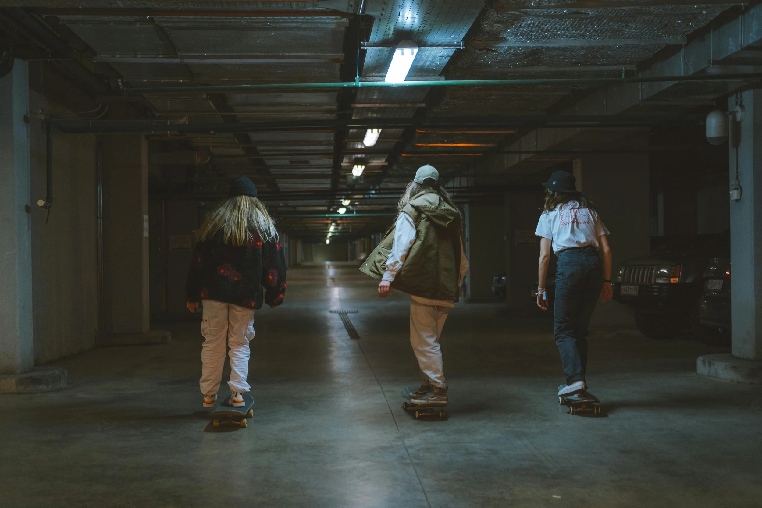 Three young women skateboarding in an underground parking garage with concrete floors, parked cars, and exposed ceiling pipes.