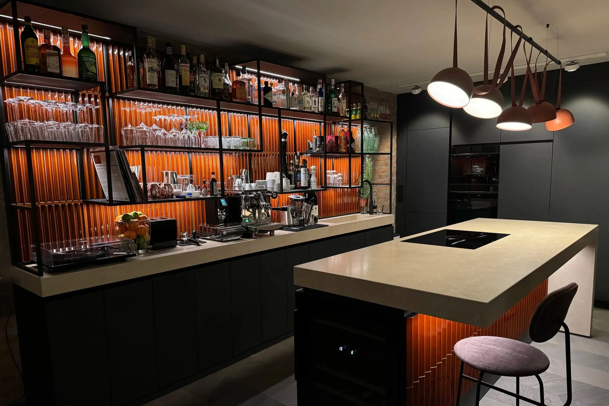 Modern bar with illuminated shelves holding bottles and glassware, a white countertop with a built-in stovetop, and a seating area with a stool, stylish hanging lights, and a dark wall background.