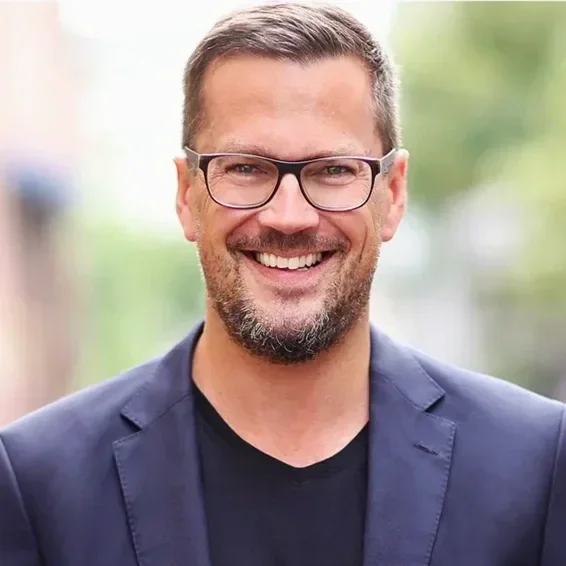 Ein lächelnder Mann mit Brille, kurzen braunen Haaren und Bart, gekleidet in einen dunklen Blazer und ein schwarzes Hemd, steht im Freien vor einem verschwommenen Hintergrund aus Bäumen und Gebäuden.