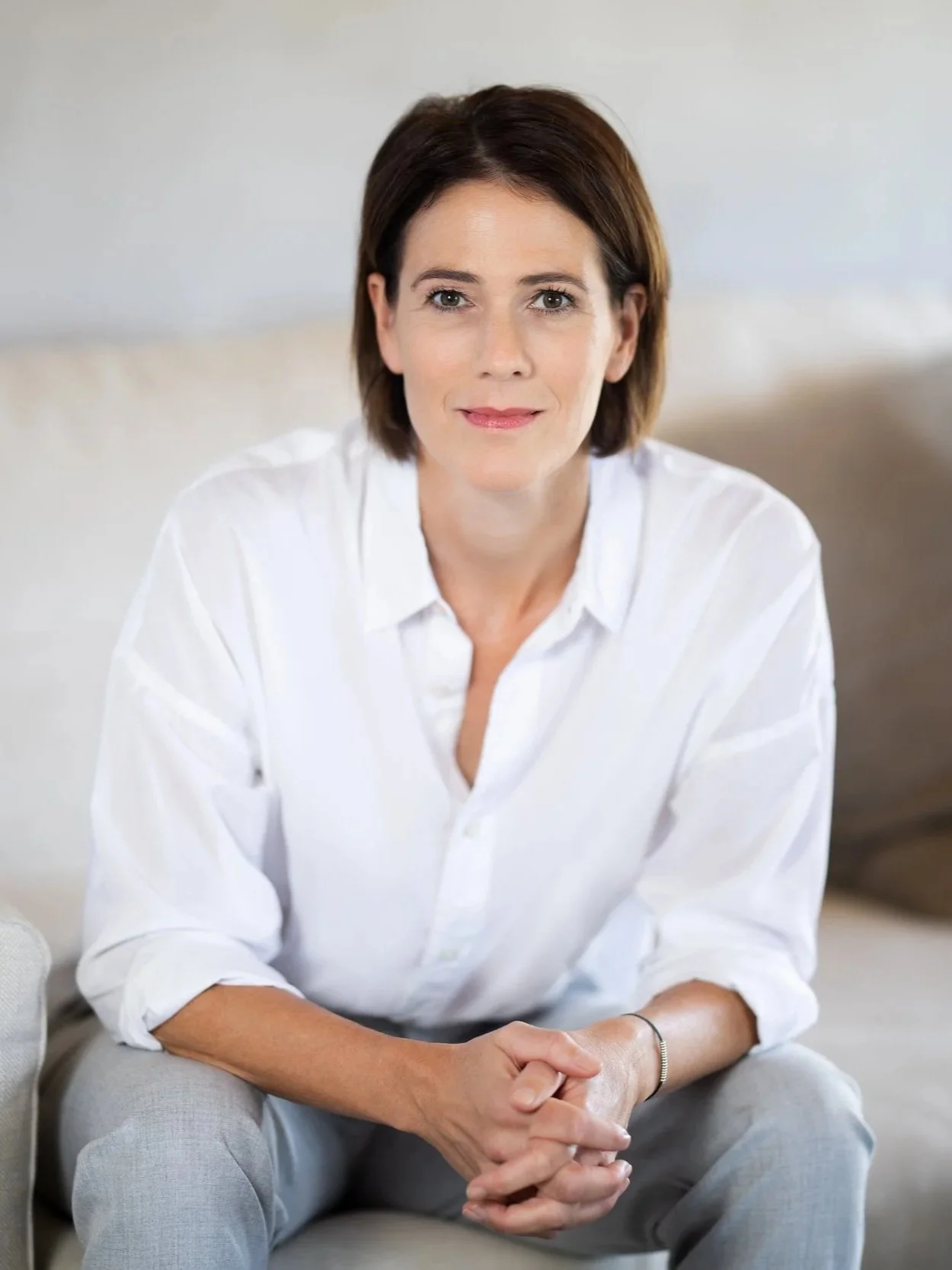 Eine Frau mit kurzen braunen Haaren und weißem Hemd sitzt auf einem beigen Sofa.