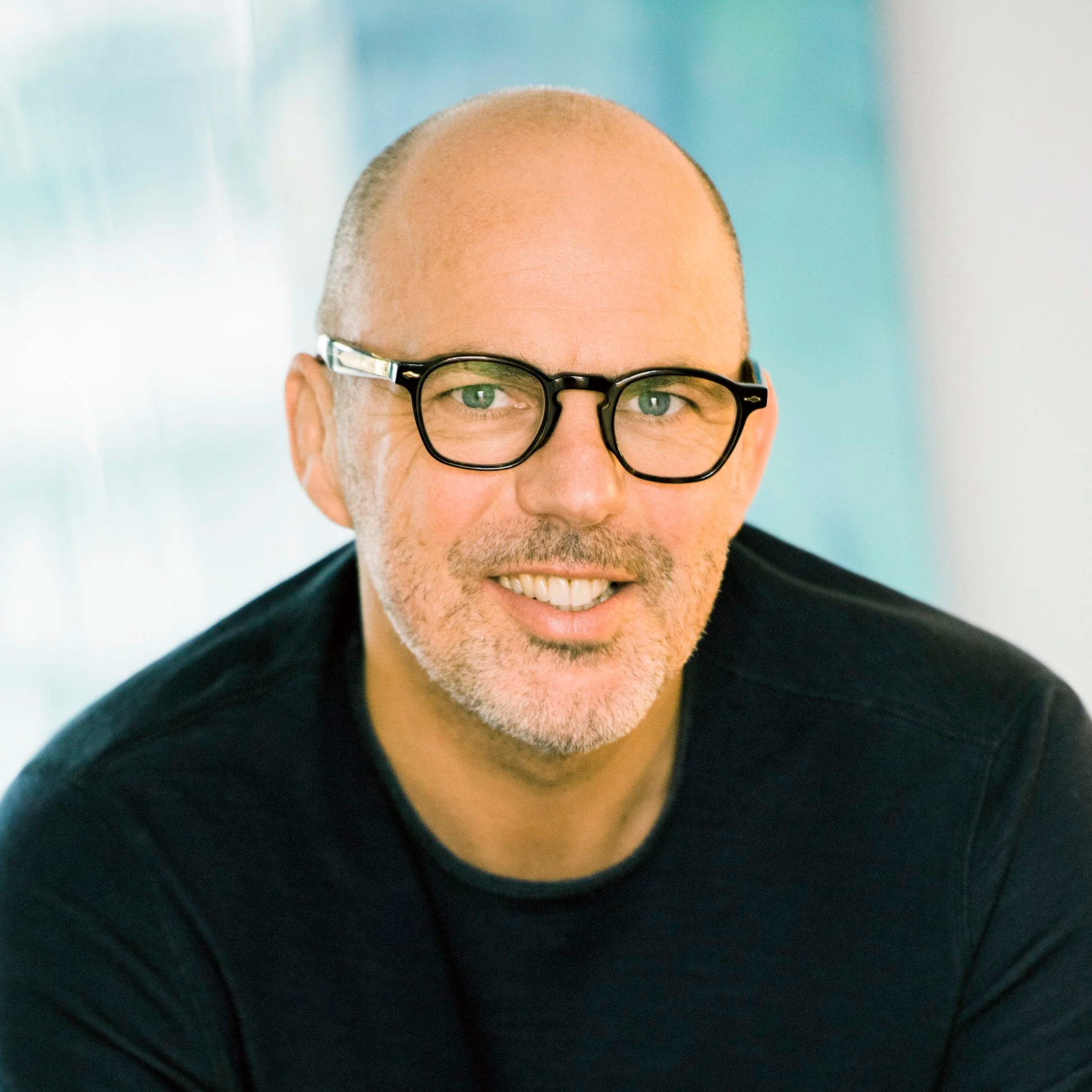 Nahaufnahme eines lächelnden Mannes mittleren Alters mit Brille, kurzem grauen Bart und rasiertem Kopf, der ein schwarzes Hemd trägt, vor einem unscharfen Hintergrund.