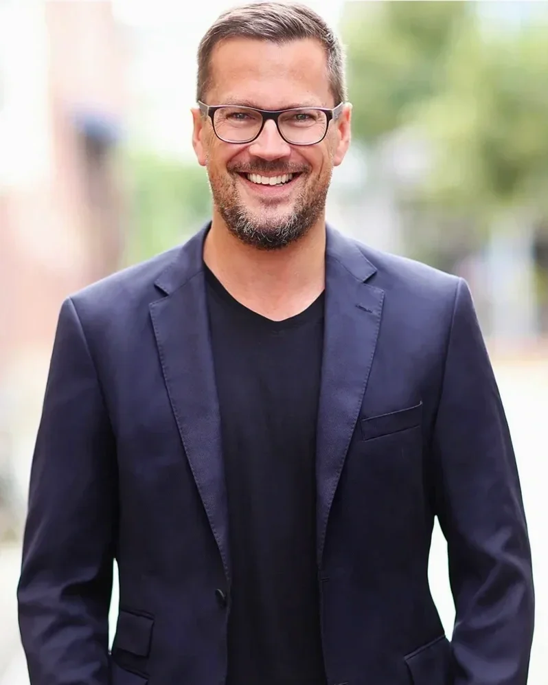 Lächelnder Mann mit Brille, dunkelblauen Blazer und schwarzem Hemd, im Freien vor unscharfem Grün im Hintergrund.