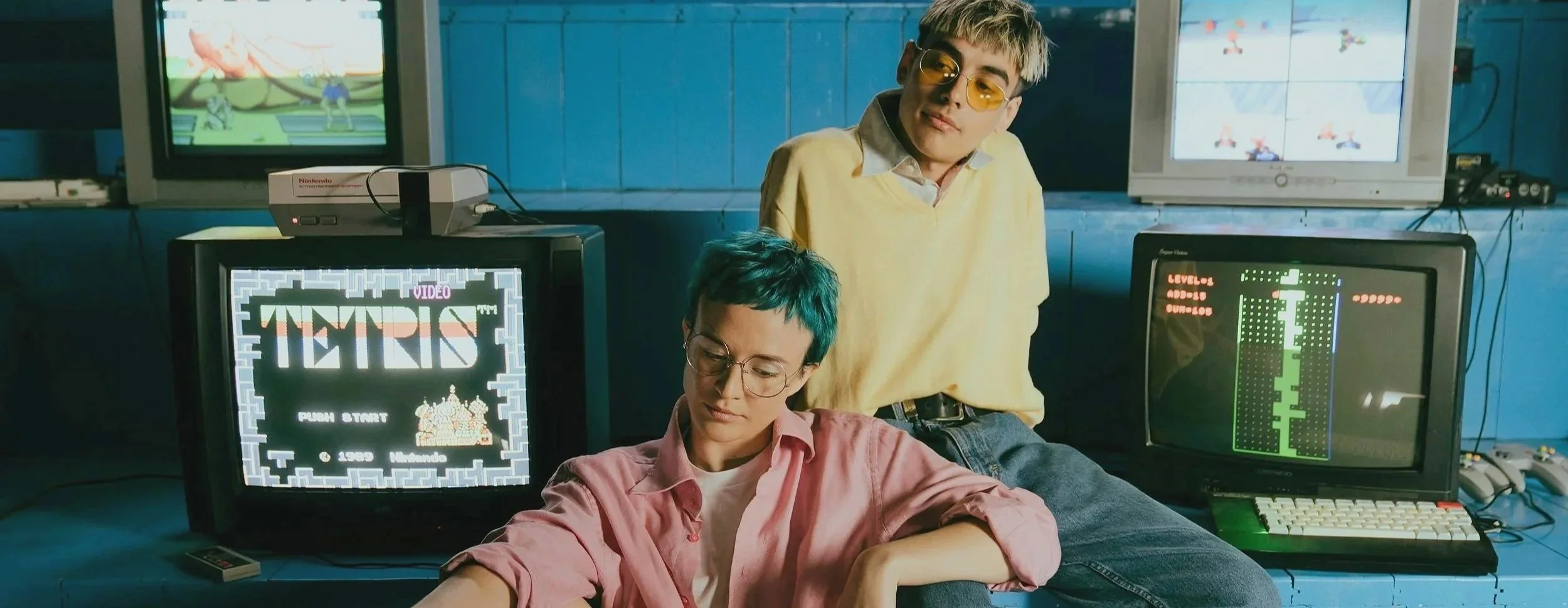 Two young women with short hair and glasses sit among vintage gaming consoles and CRT televisions playing Tetris. One woman has turquoise hair and is wearing a pink shirt, while the other has blonde hair with dark roots and a yellow sweater.