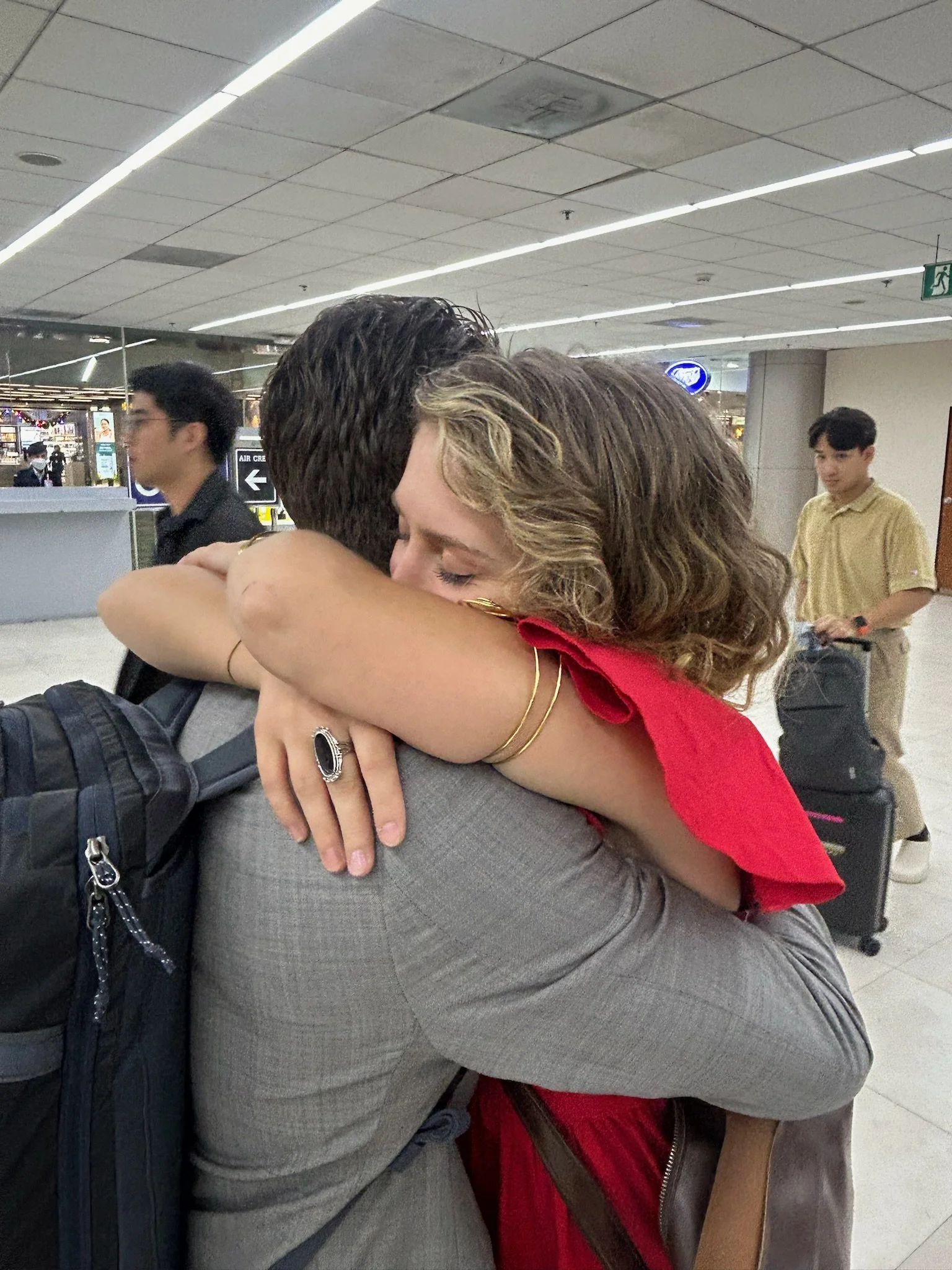 Two people hugging at an airport, with luggage and other travelers in the background.