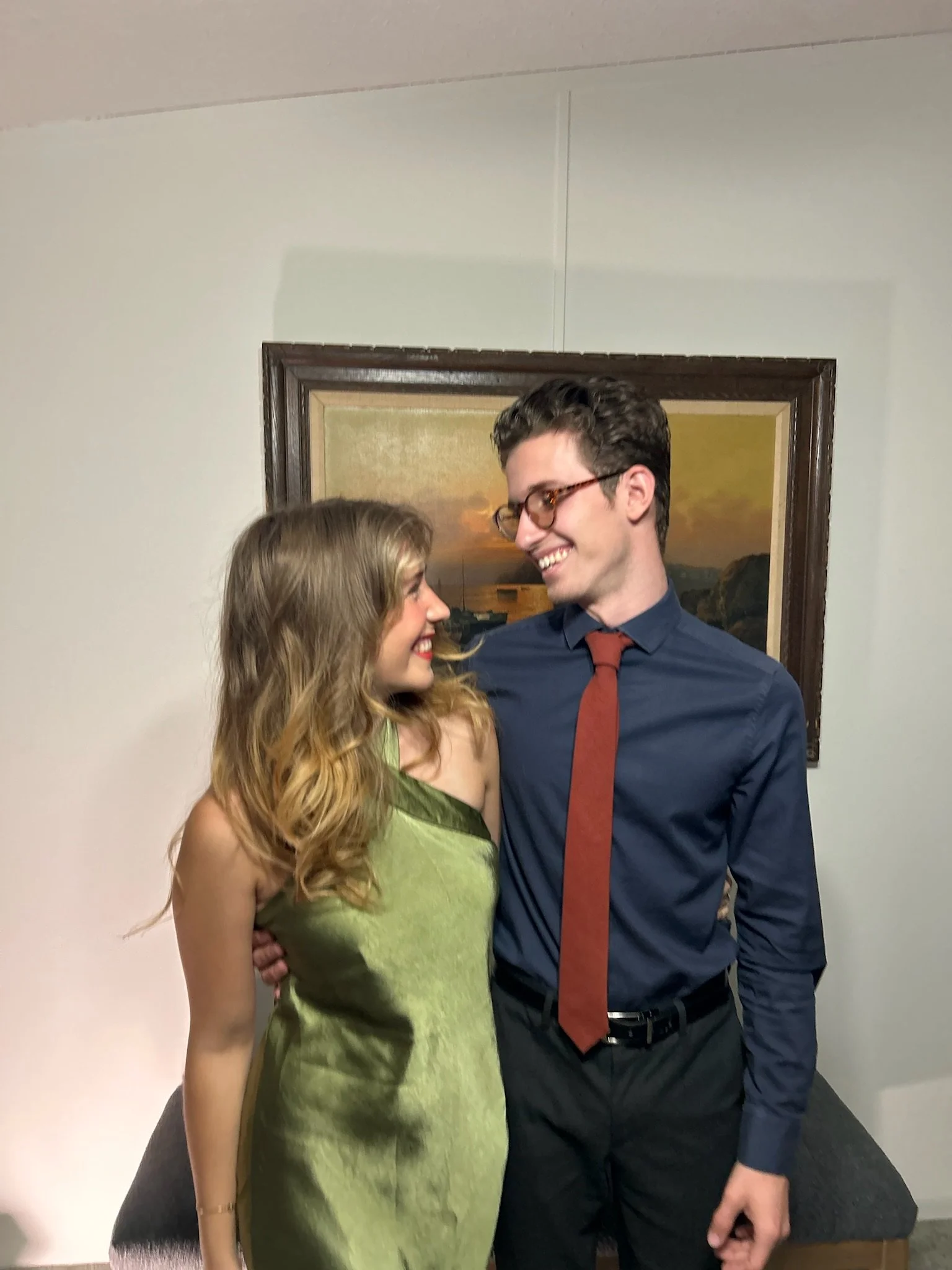 A man and woman standing close together, smiling and looking at each other indoors in front of a framed painting. The woman has long, wavy blonde hair and is wearing a sleeveless green dress. The man has short, dark hair, glasses, and is dressed in a